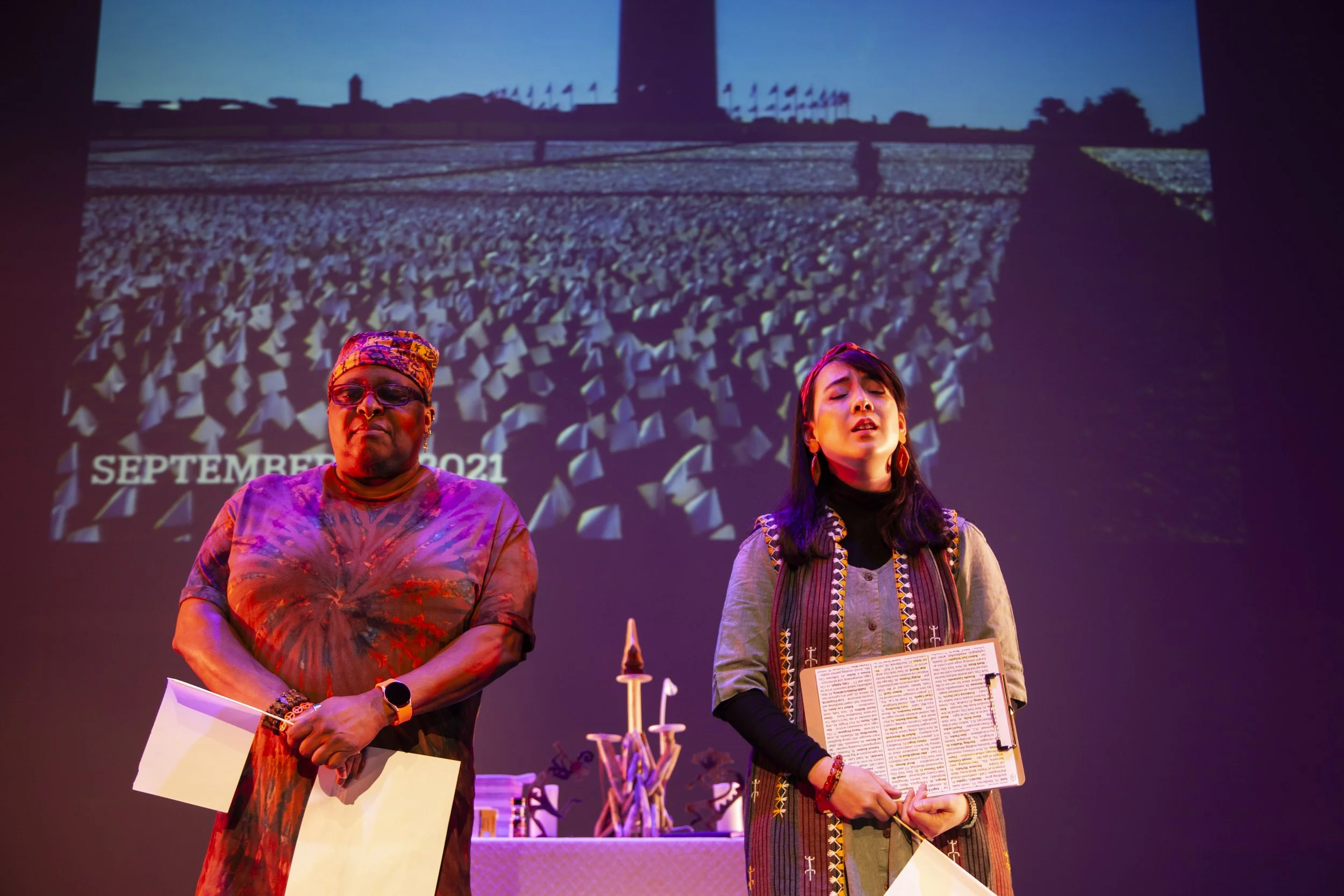 Two women standing on stage during a performance or event, with a large screen behind them displaying an image of a field with flags, a tower, and the text 'September 2021'.
