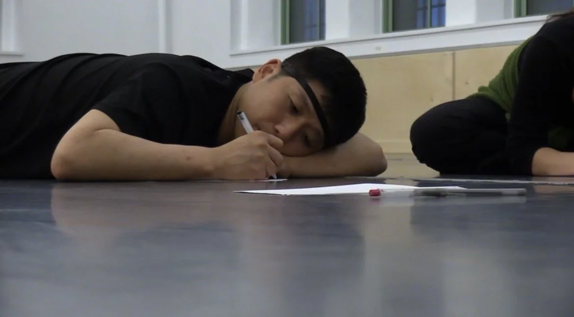 A person lying on the floor, drawing or writing on a piece of paper, with a marker and eraser nearby.