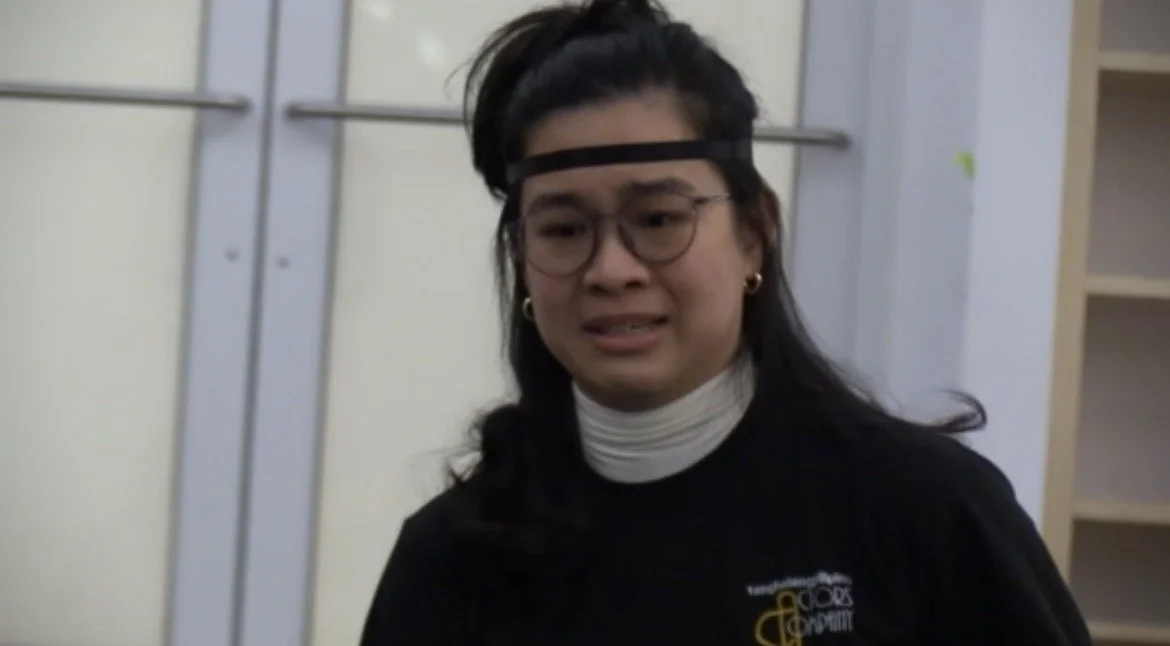 A woman with glasses and long dark hair wearing a black t-shirt, white turtleneck, and headband, appears upset or crying, standing in front of a light-colored wall with a metal door or pipe visible in the background.