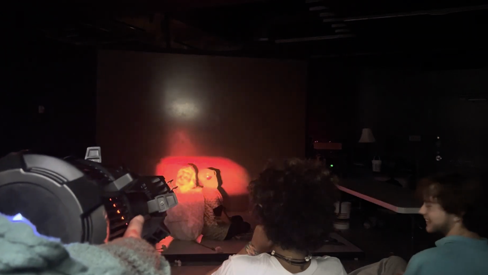 Two people sitting on a table in a dark room watching a shadow puppet show projected on the wall, with a person operating a flashlight or projector in the foreground.