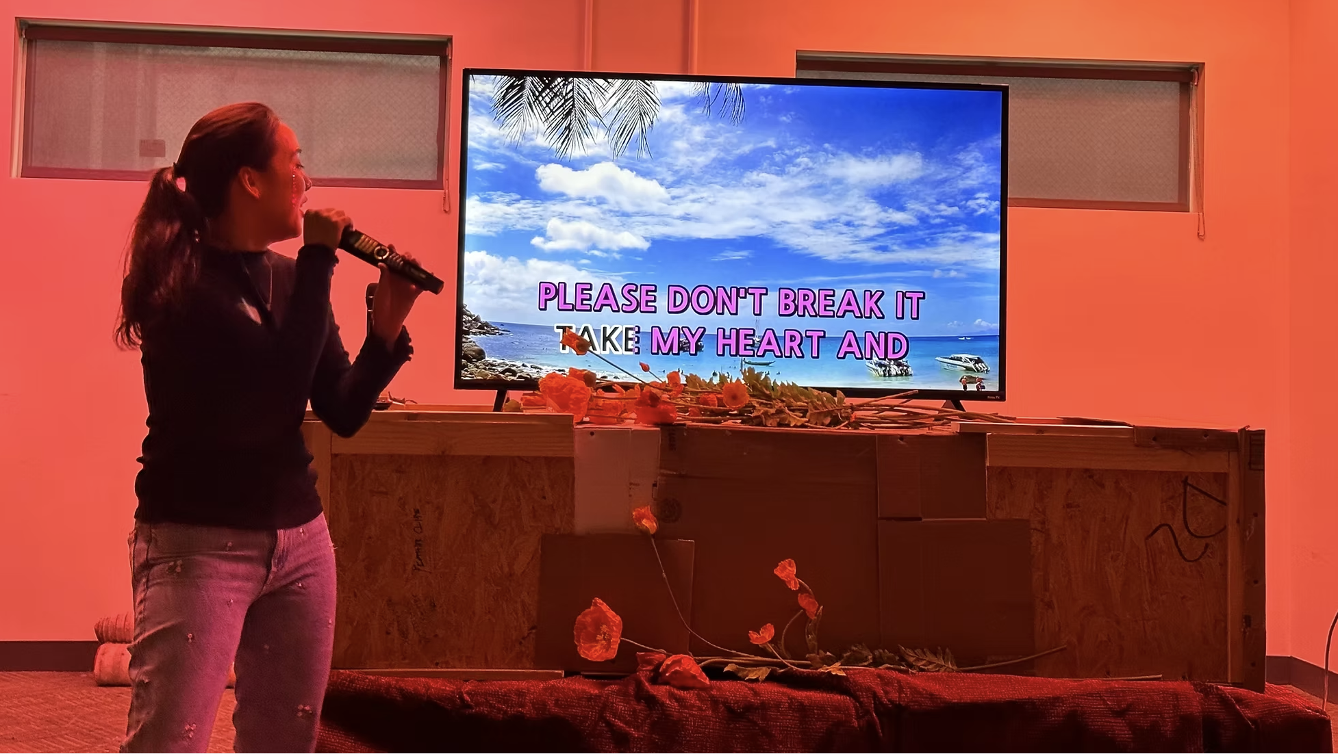 A woman singing into a microphone with a TV behind her displaying a beach scene and song lyrics reading "Please don't break it, take my heart and". Flowers are arranged on a wooden surface in front of the TV.