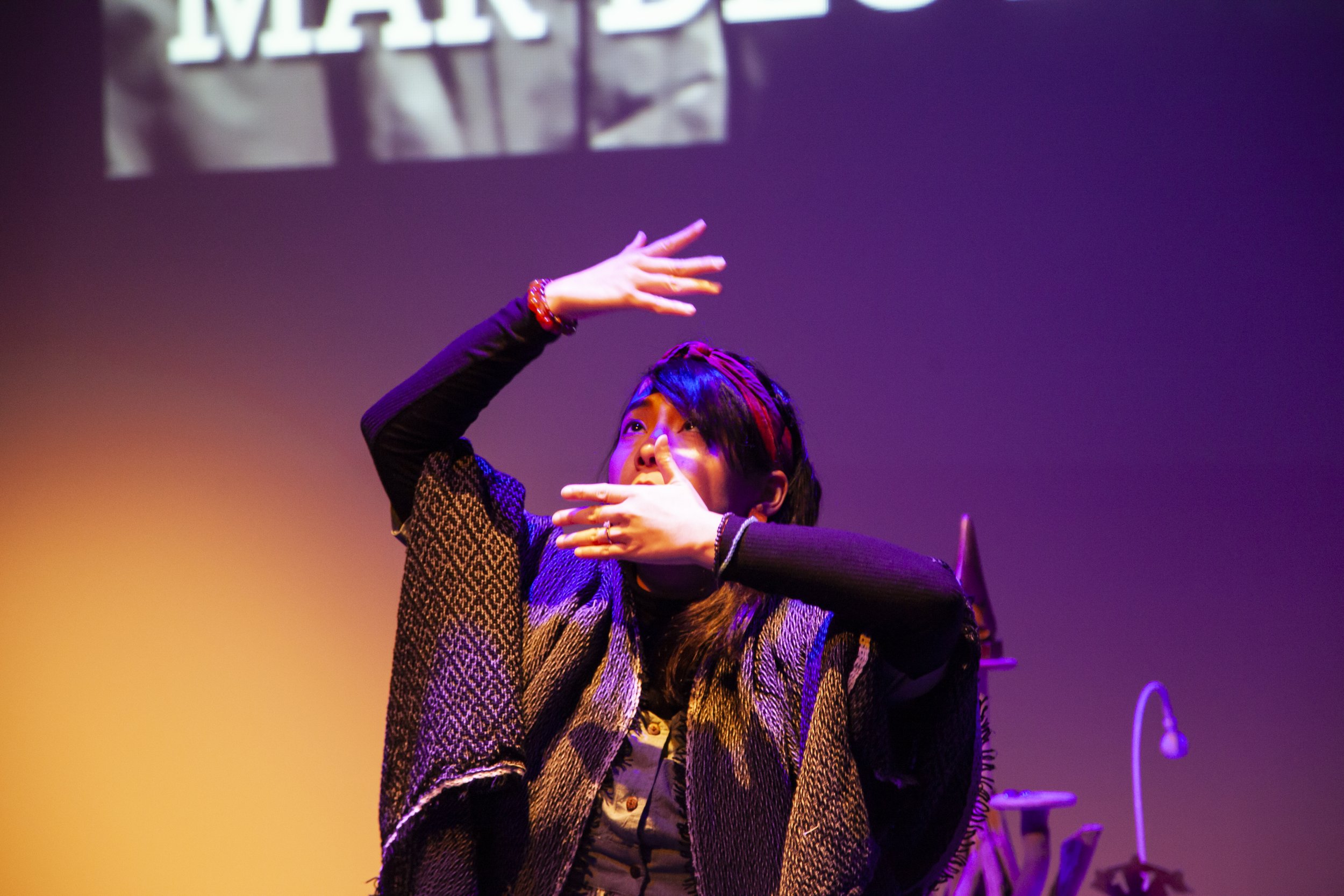A person with dark hair, wearing a patterned shawl, performing a dance move on a stage with purple and yellow lighting, with part of a sign visible in the background.