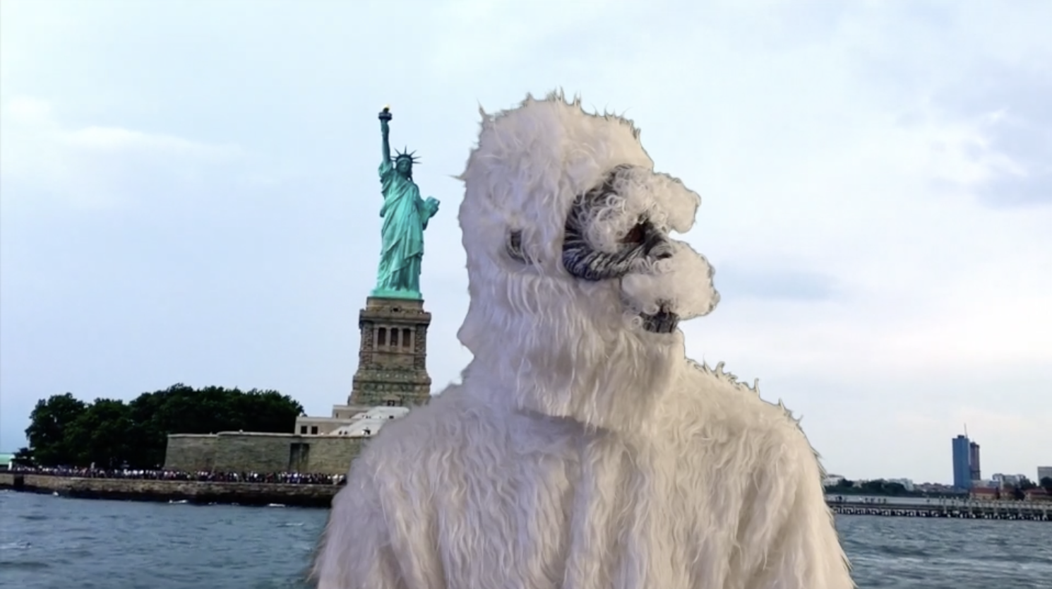 A giant white fluffy dog with a beard standing in front of the Statue of Liberty in New York City.