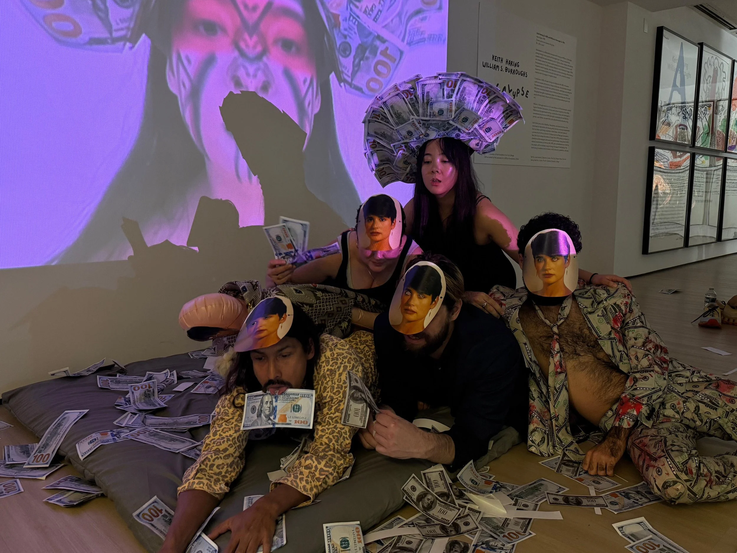 Group of five people lounging on a bed covered with paper money, with cardboard cutouts of a woman wearing a pink mask and a man with a dollar bill face mask. Two individuals are wearing masks with a woman's face on them. One person has a large headpiece made of dollar bills, and a large projection of a woman's face with face paint and dollar bills behind them is in the background. The scene appears to be an art installation.