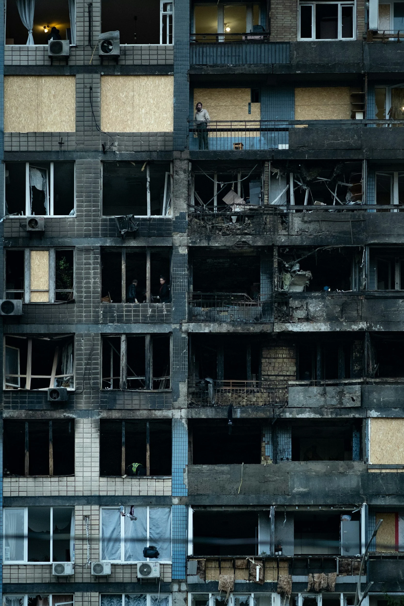 The building's facade is heavily damaged with broken and boarded-up windows, charred walls, and missing sections, indicating fire damage. Two people are seen on a balcony, one man standing and looking outward, the other person inspecting the damage inside. The overall scene suggests aftermath of a fire or major destructive event.