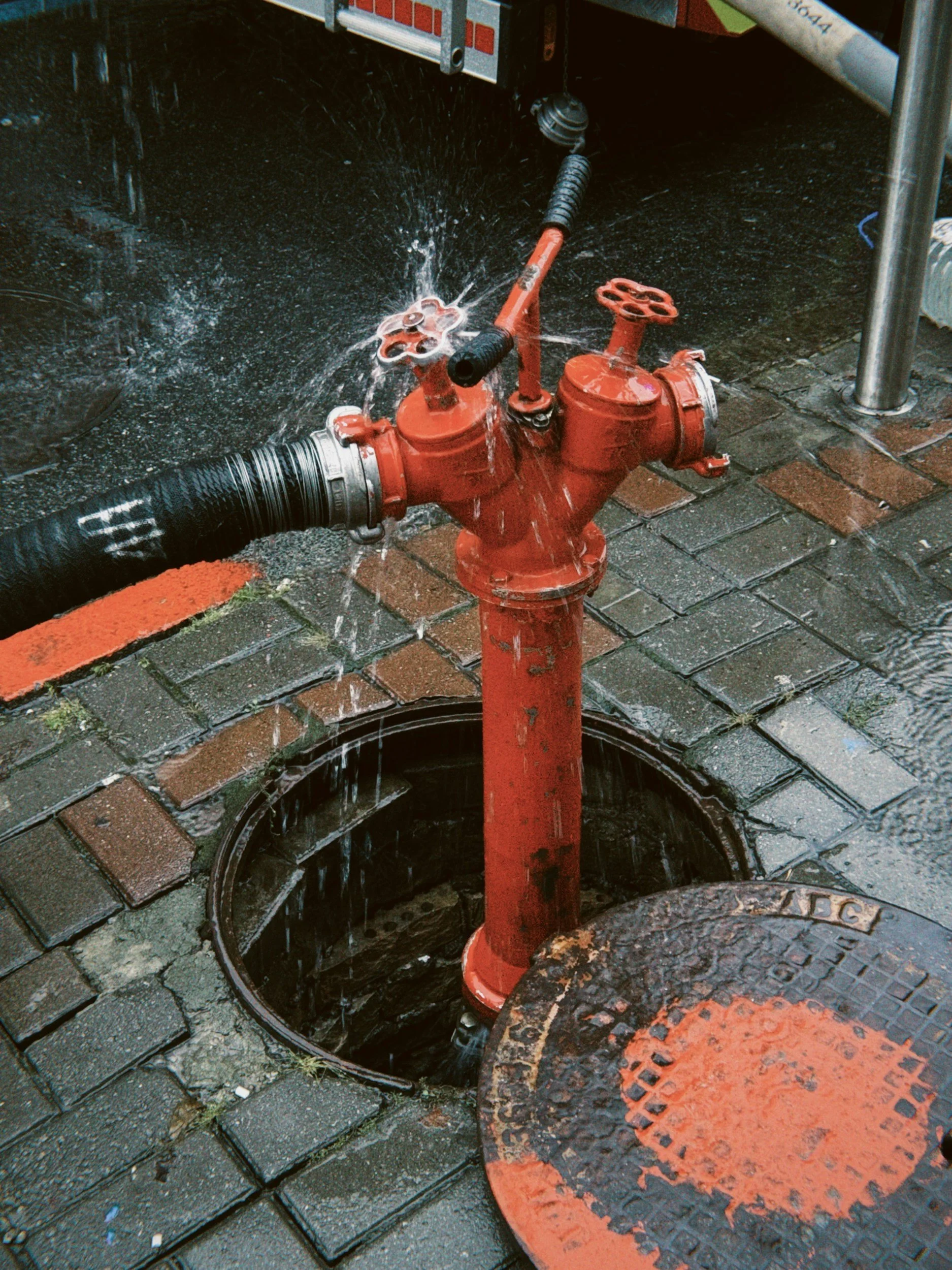 A red fire hydrant on a brick sidewalk, with water spraying from two open valves, and a round, rusted metal cover nearby.