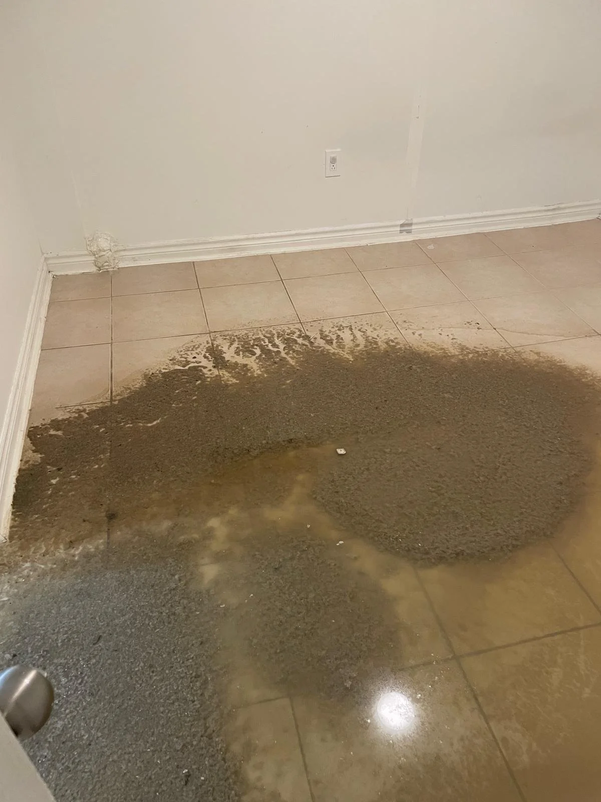 A tiled floor shows a large wet brown stain near a white wall with a power outlet and baseboard molding.