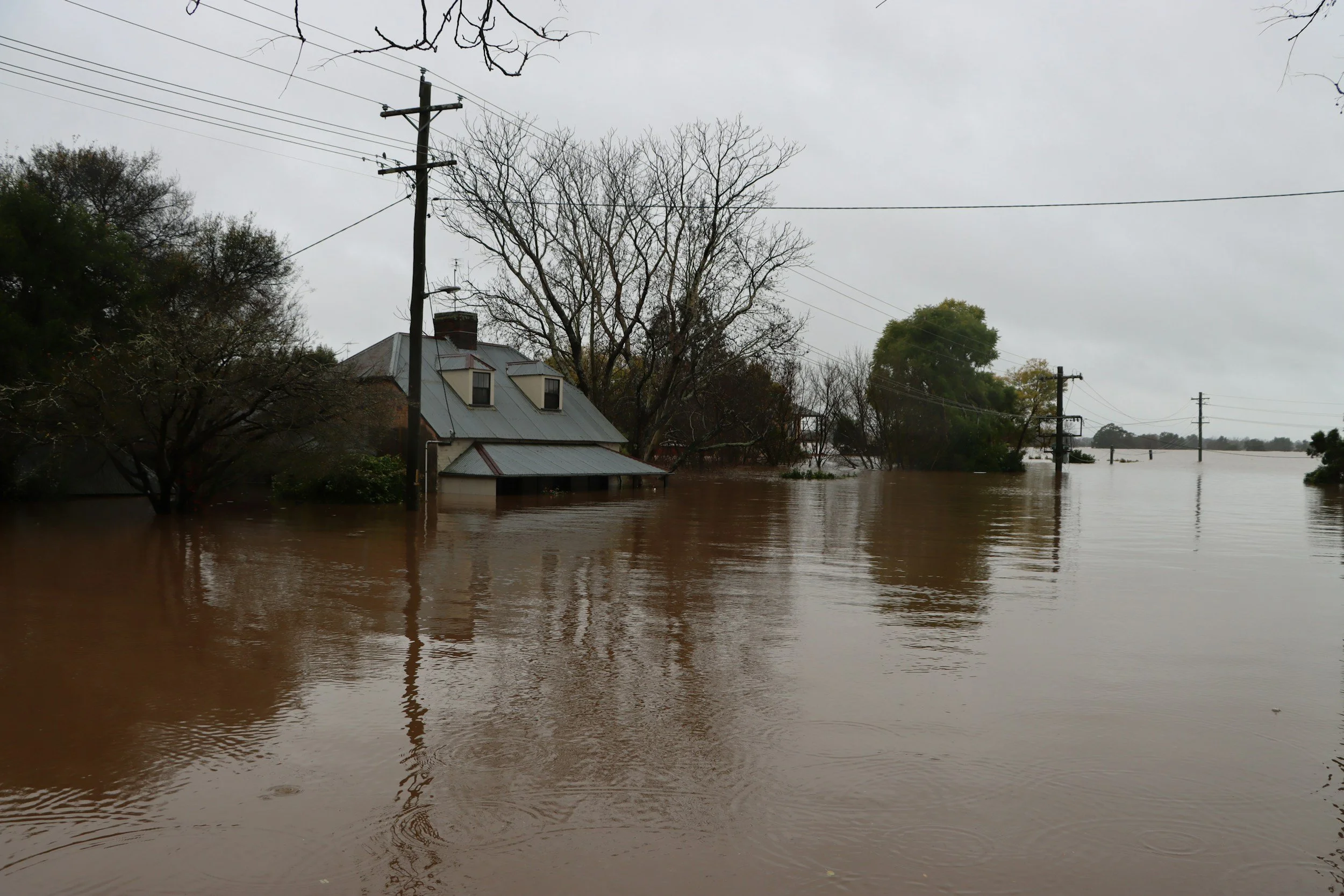 A house and power lines are partially submerged in floodwaters, with trees in the background and an overcast sky.