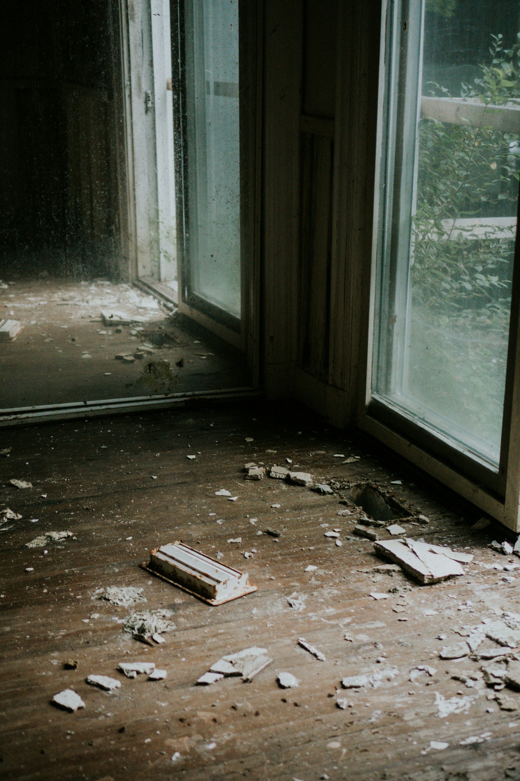 Interior of a dilapidated room with a sliding glass door leading outside, scattered debris and broken pieces of drywall on the wooden floor, with some plants visible outside through the glass.