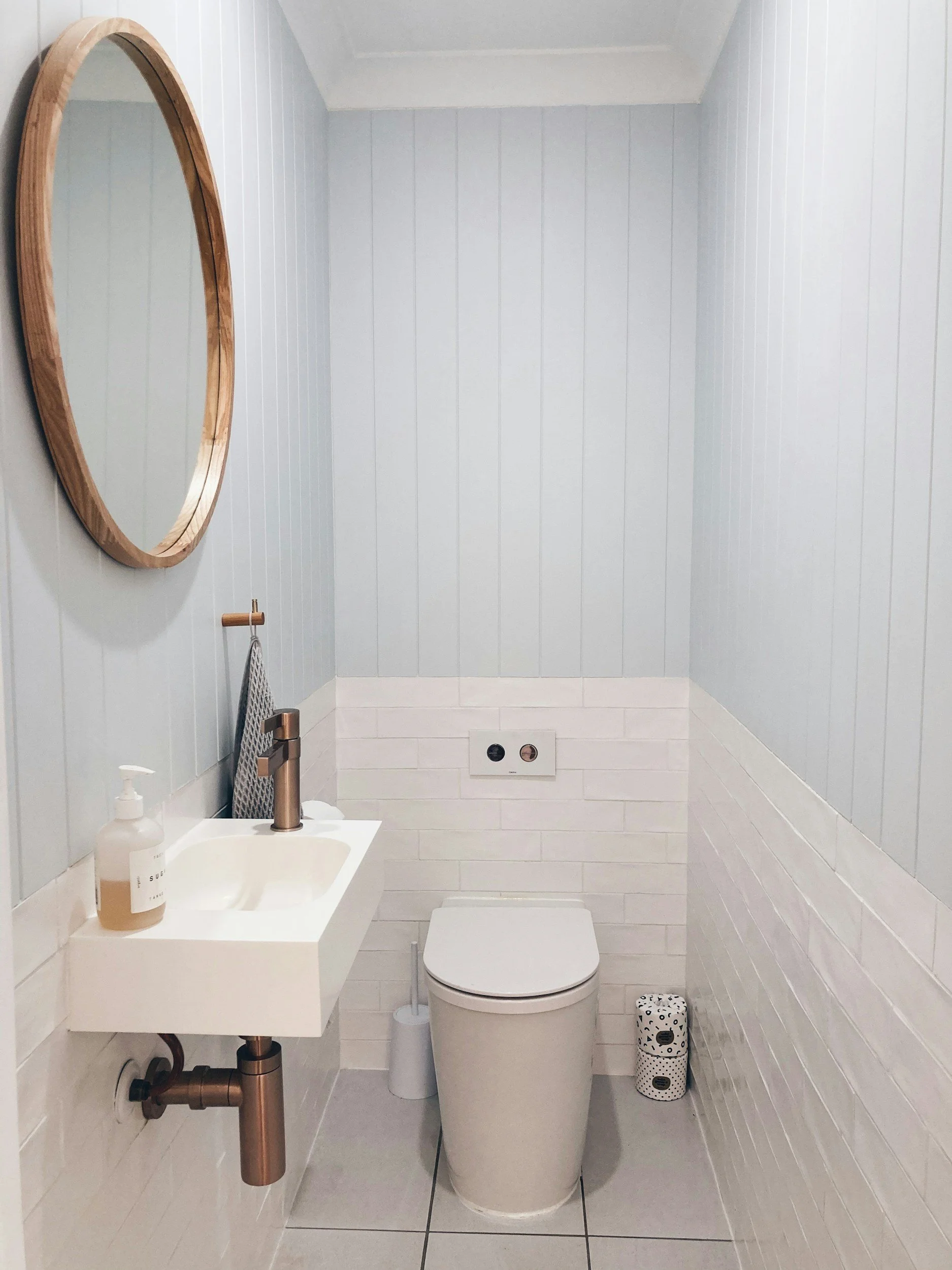 Small bathroom with light blue panel walls, white tiling, small white sink with copper fittings, and a modern white toilet. Small decorative toilet paper holder and a hand soap dispenser on the sink, oval mirror on the wall.