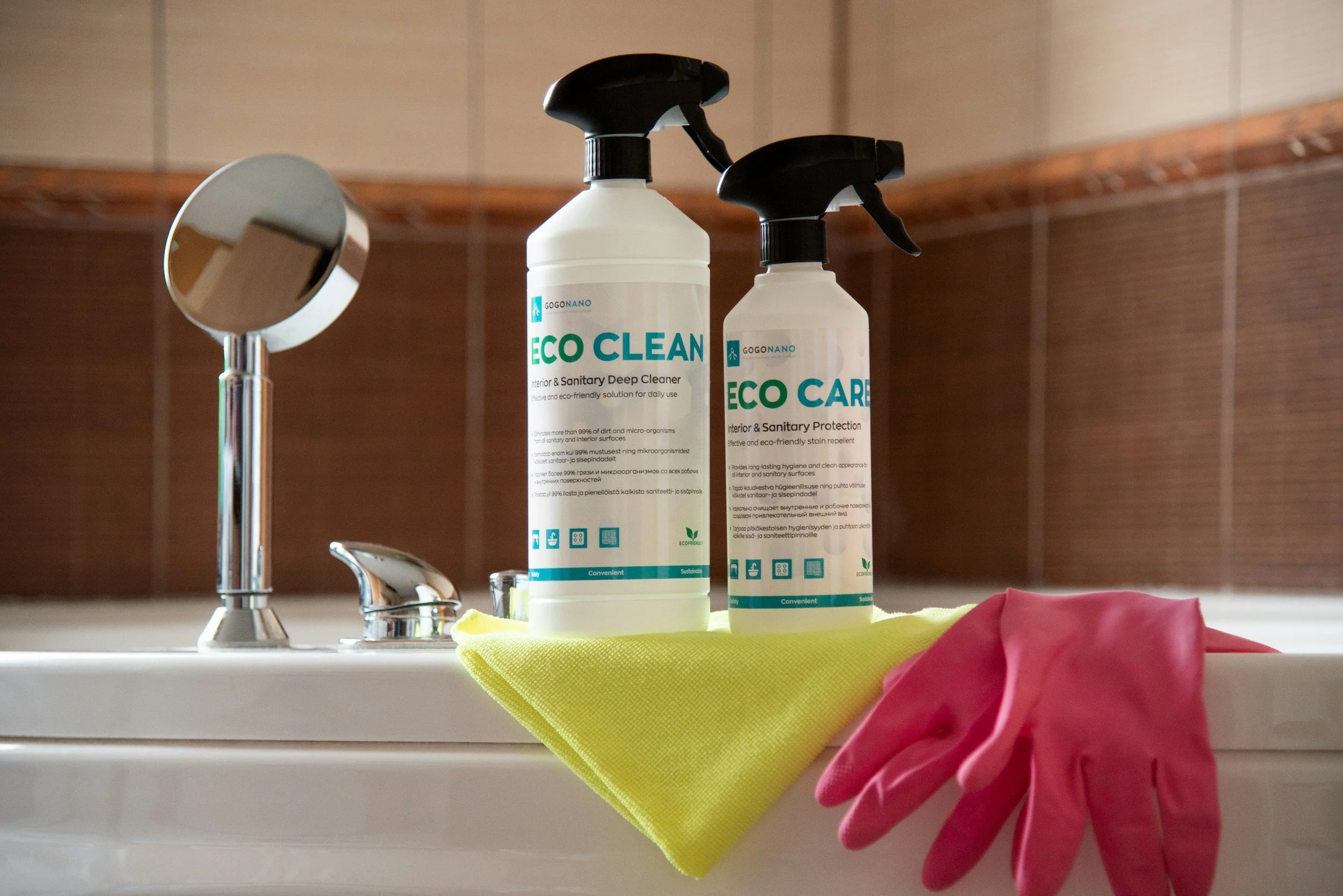 Cleaning supplies on a bathroom sink, including bottles of Eco Clean, pink rubber gloves, and a yellow cloth, with a mirror and faucet in the background.
