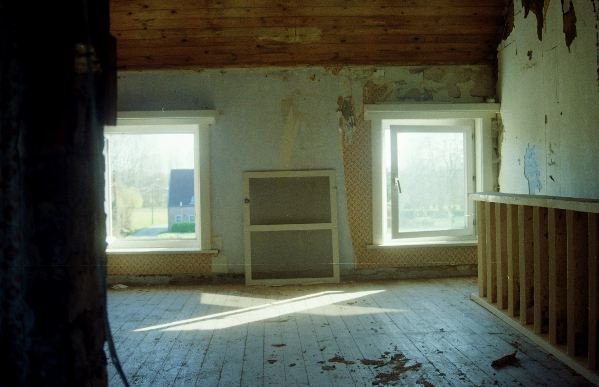 Empty room in a house under renovation with two windows, partially exposed wall, and a wooden floor with dirt and debris.