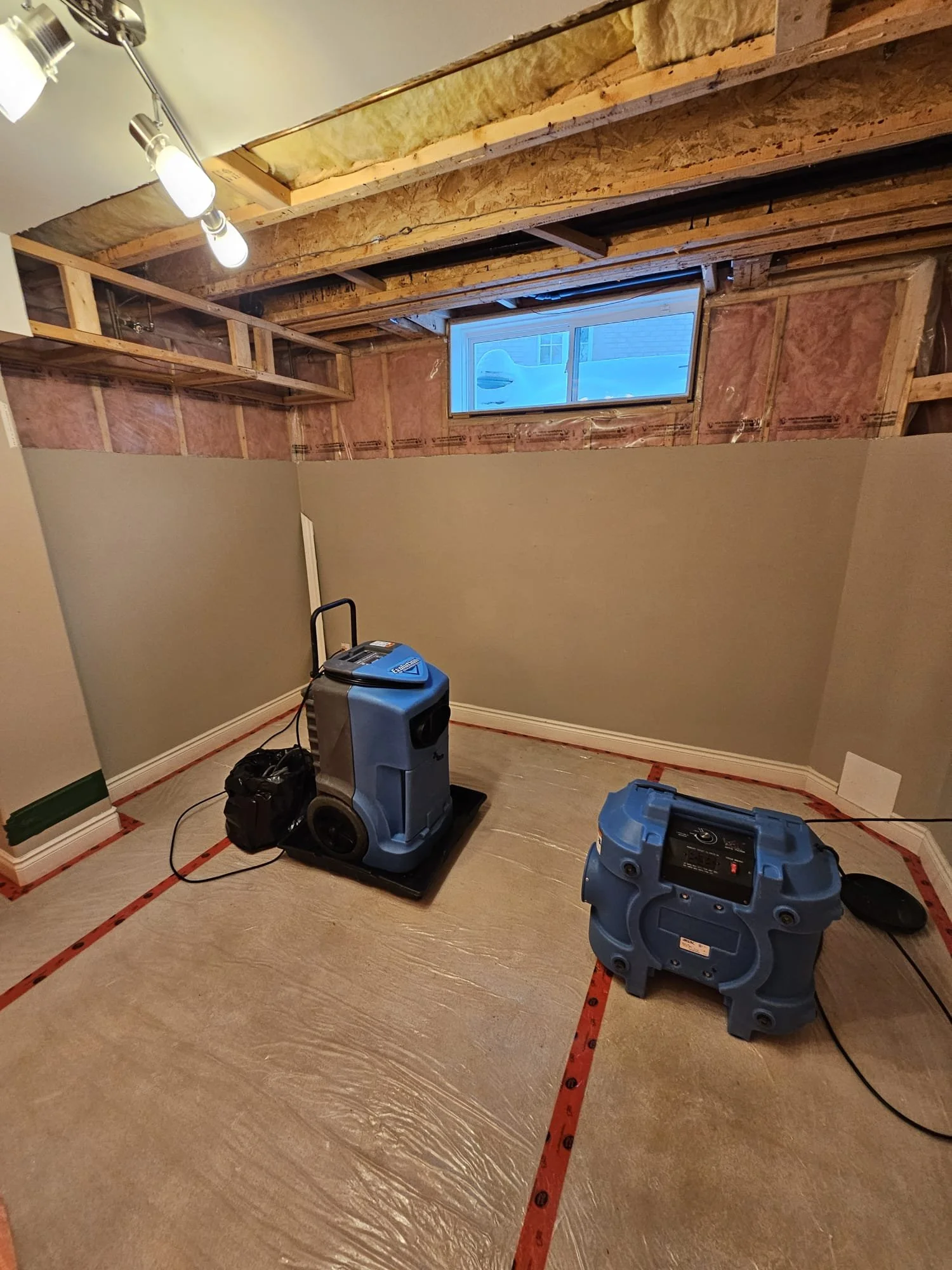 A partially finished basement room under construction with insulated ceiling and a small high window. The floor is covered with plastic sheeting, and there are two large air scrubbers or vacuums in the room.