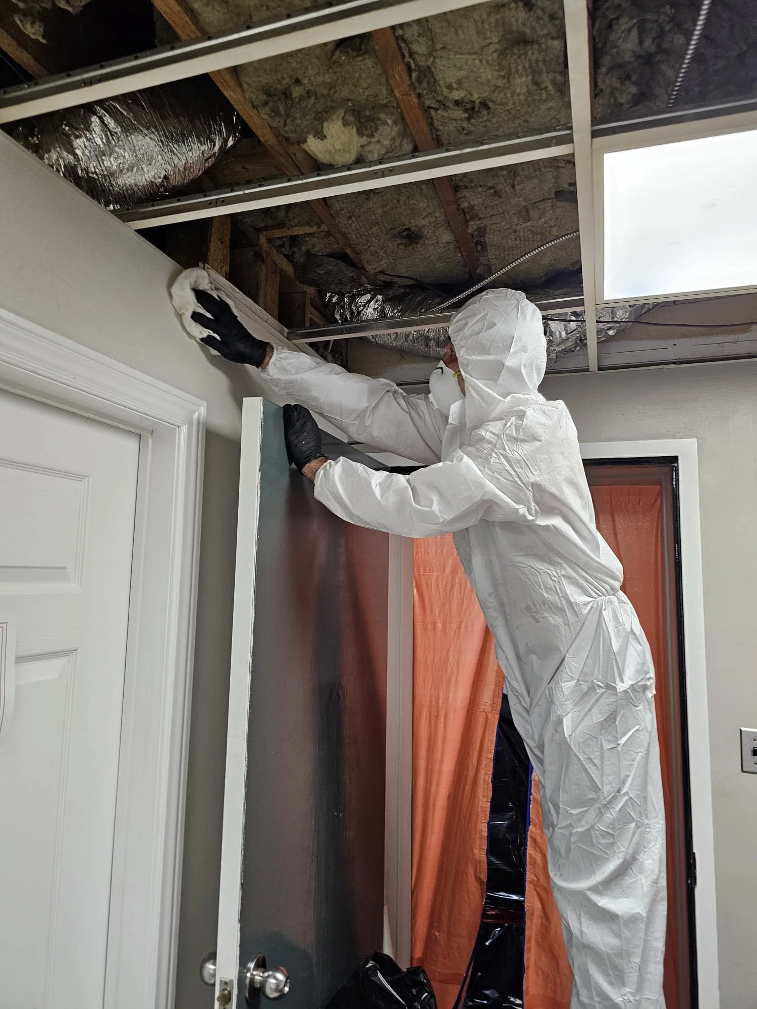 A person wearing white protective coveralls, black gloves, and a face mask, installing a door or panel inside a room with exposed ceiling insulation.