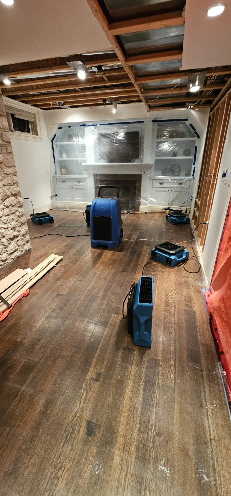 Living room undergoing renovation with floor fans and ceiling exposed, plastic sheeting on the walls, and a partially installed fireplace with built-in shelves.