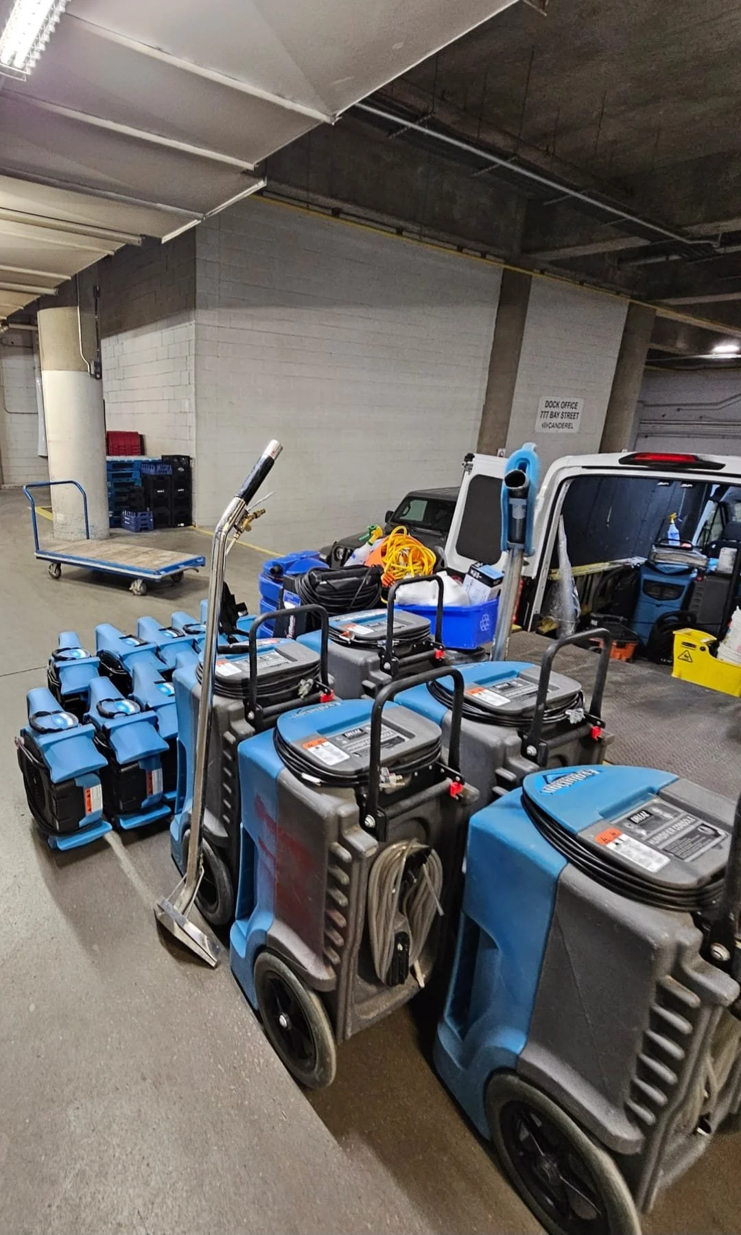 Cleaning equipment including floor scrubbers and vacuum cleaners in a parking garage, with a hand truck in the background