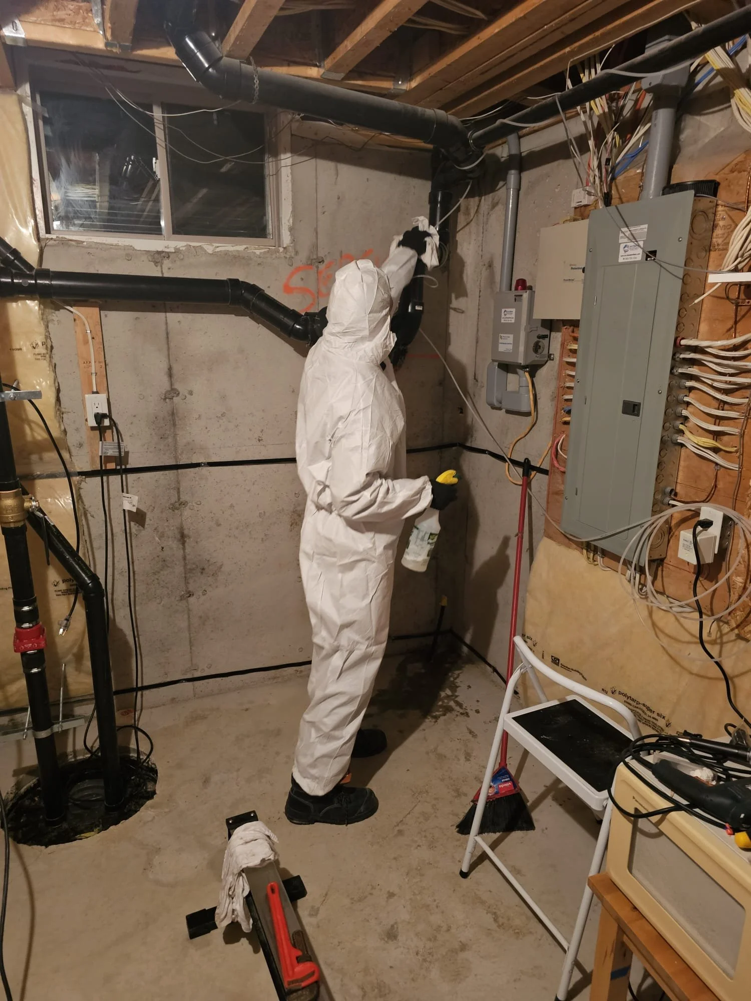 Person wearing white protective suit and gloves working on pipes in a basement or utility room.