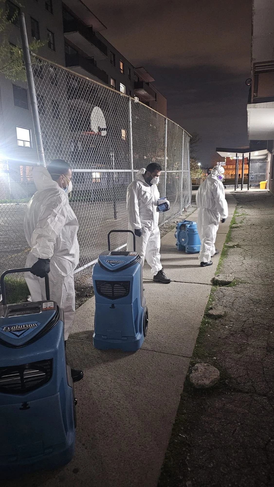 Three people dressed in white protective suits and masks are disinfecting a sidewalk at night with blue disinfectant sprayers.