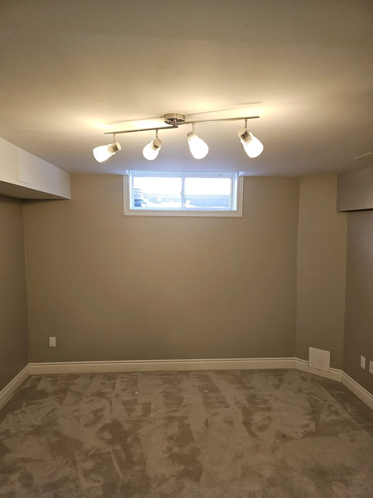 Empty room with beige walls, a small window near the ceiling, and a ceiling light fixture with four bulbs.