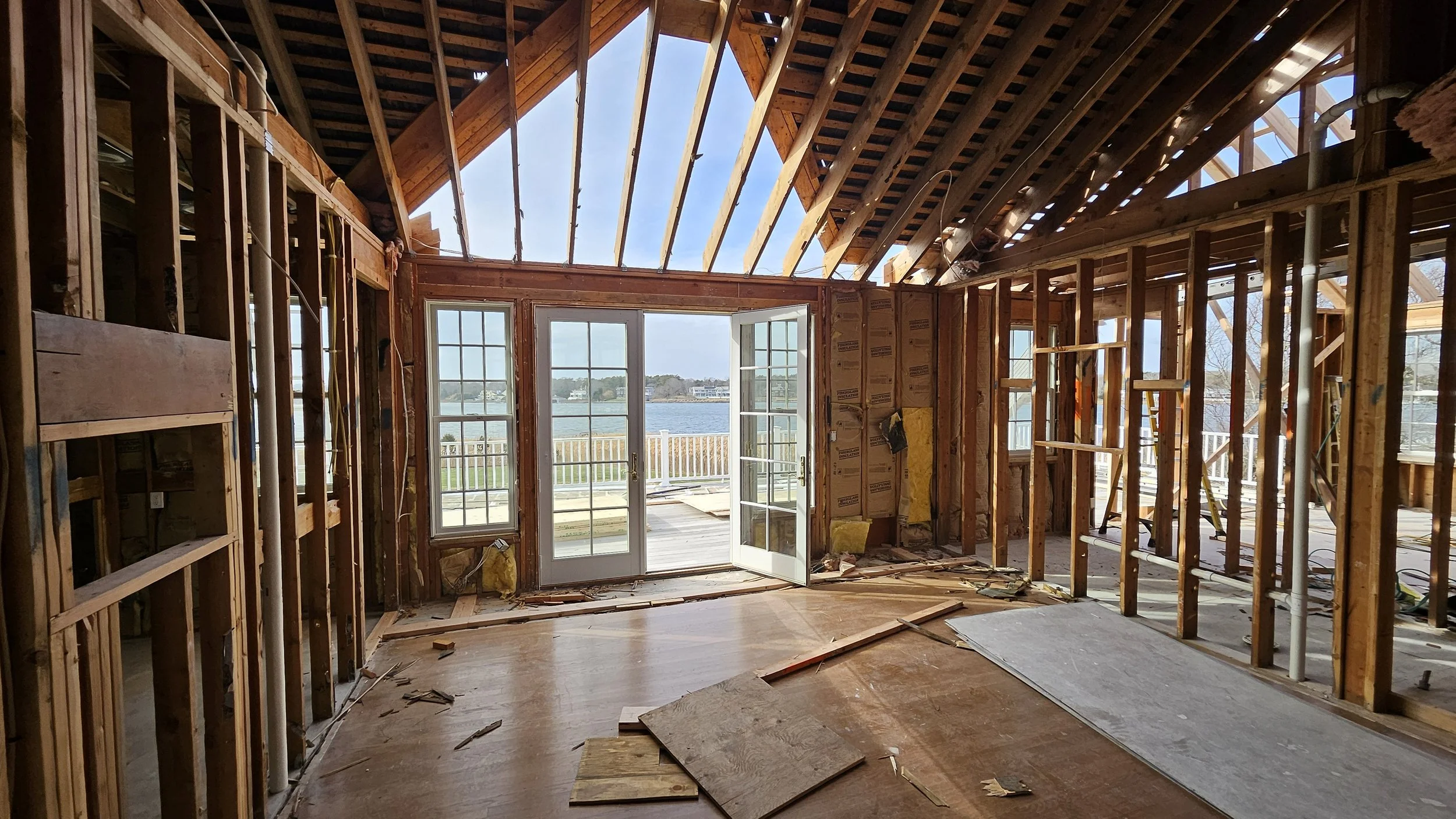 Interior of a house under construction with exposed wooden framing and an open doorway leading to a balcony overlooking a body of water.