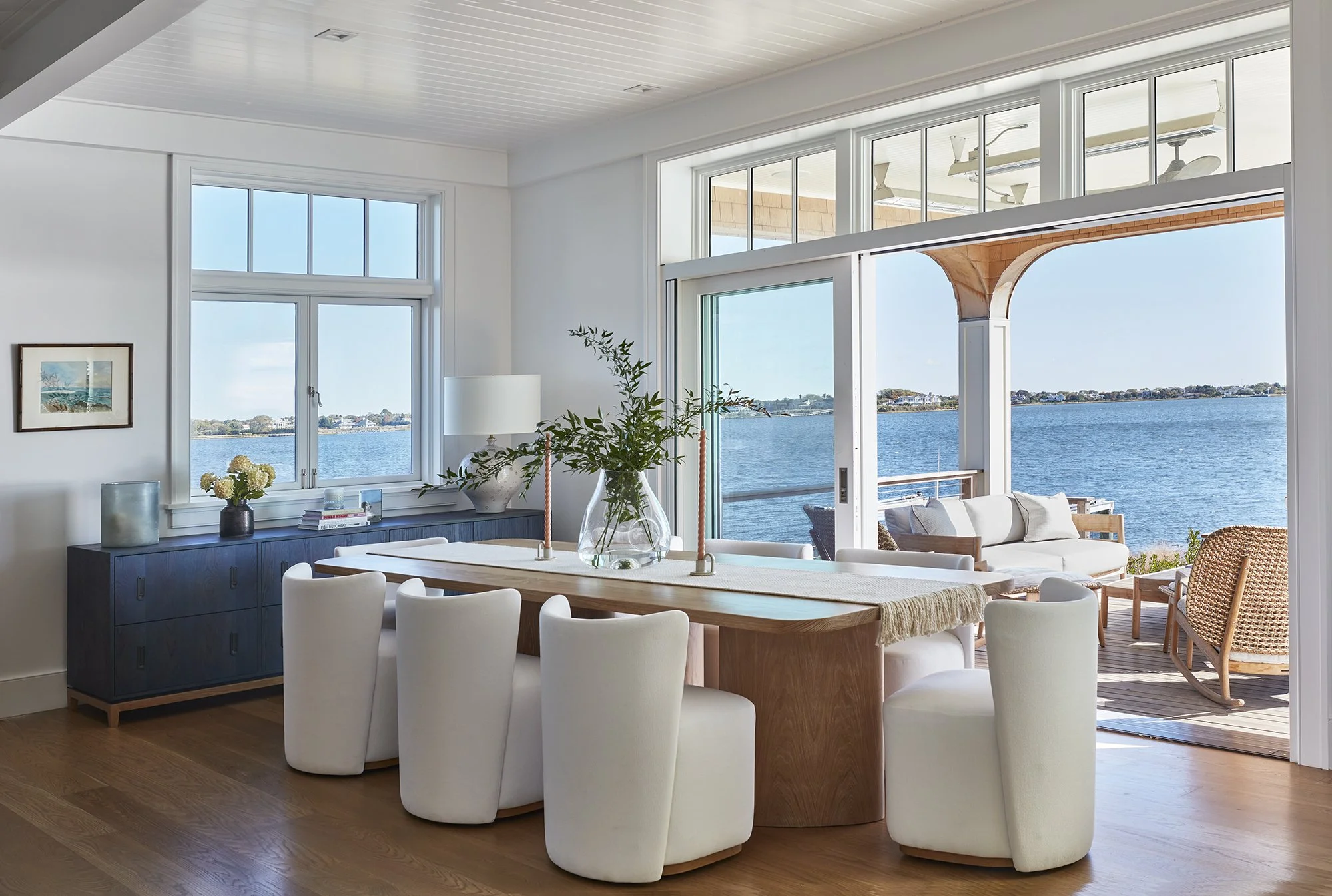 Coastal living room with large view of water, featuring a dining table surrounded by white chairs, a blue sideboard, and an outdoor patio with seating, all with generous windows and sliding door.