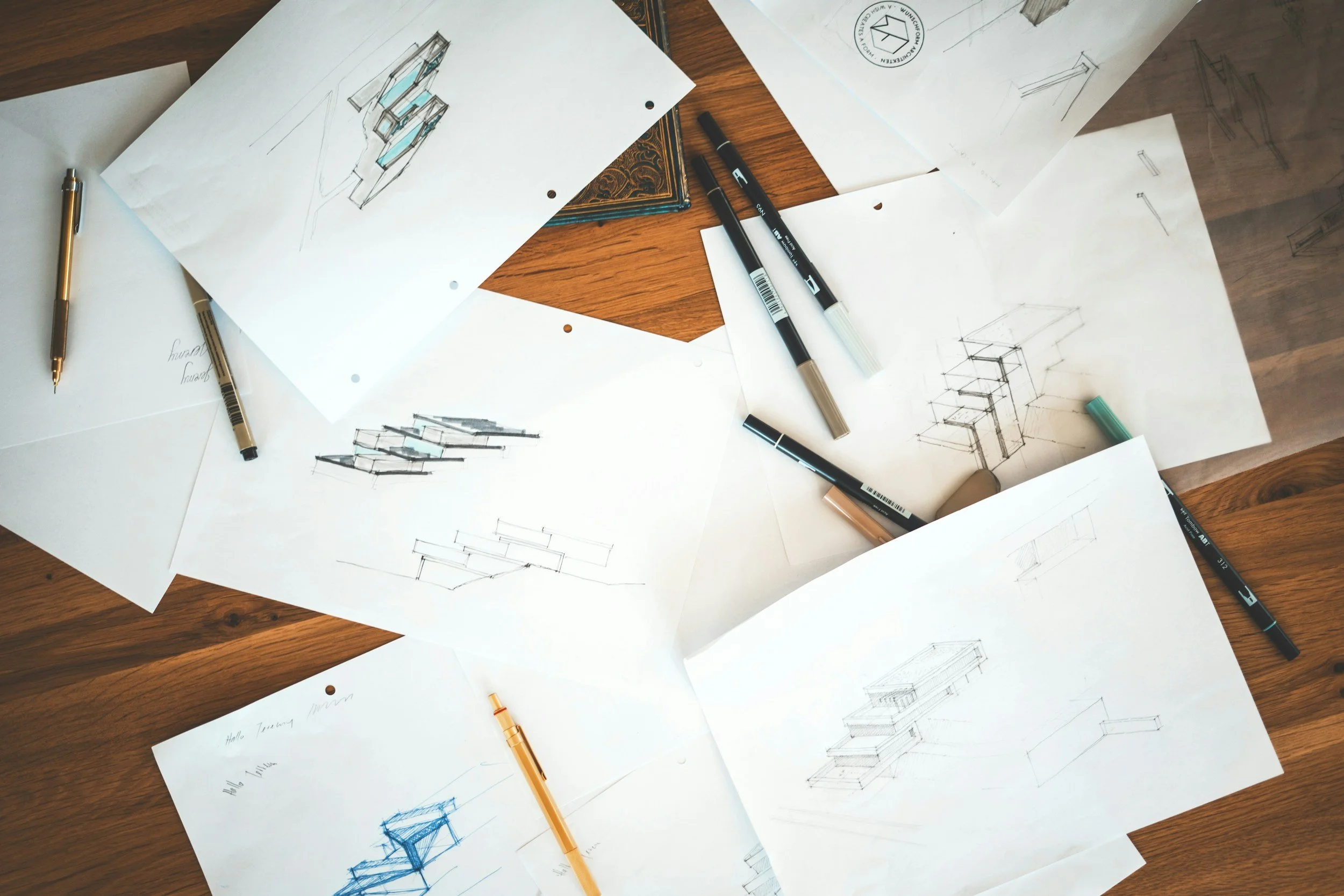 Architectural design sketches and technical drawings spread out on a wooden table with pens and markers.