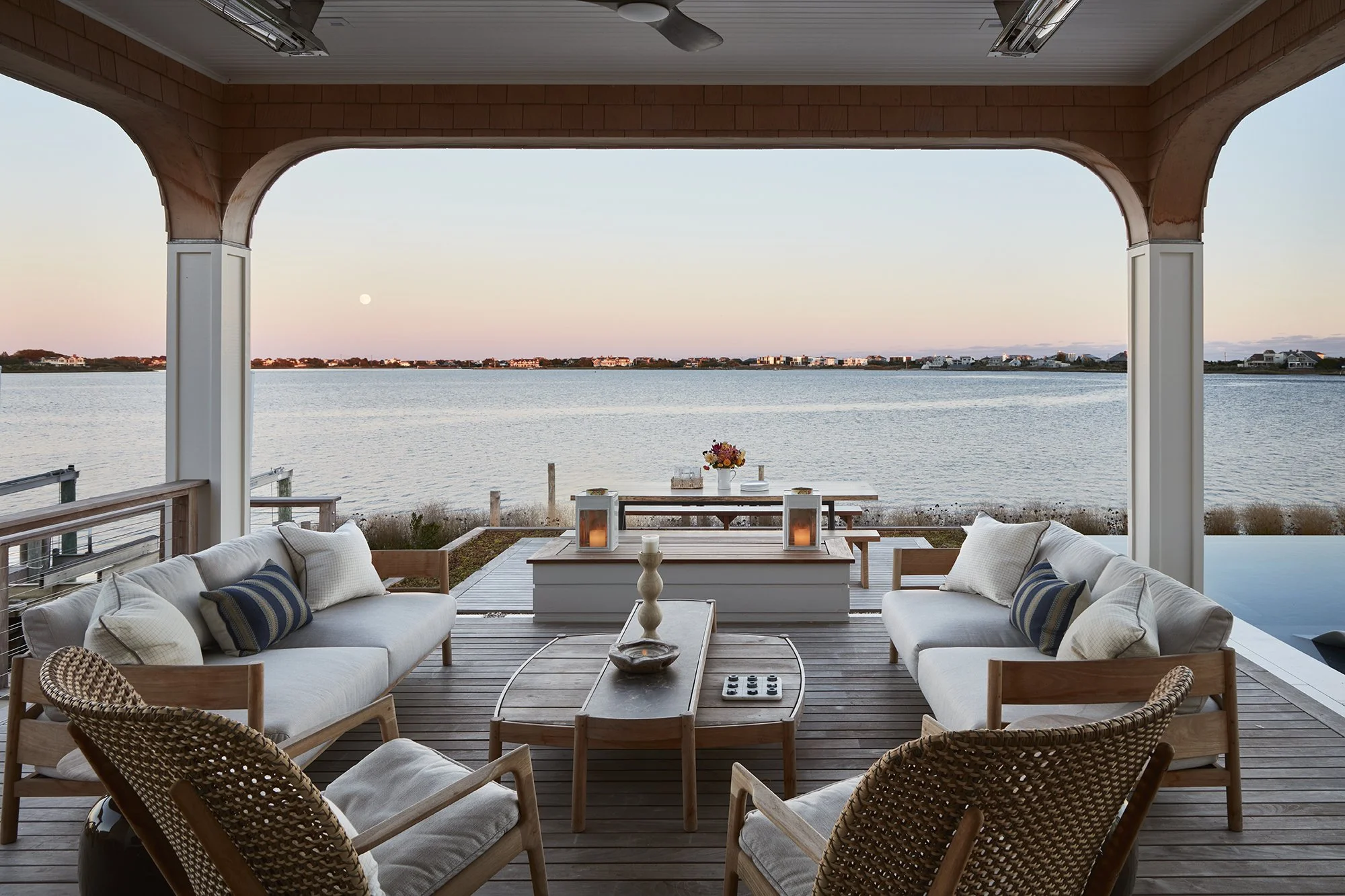 Outdoor patio overlooking water with furniture including two sofas, two armchairs, a coffee table, and a table with a vase of flowers, lanterns, and a candle, during sunset.