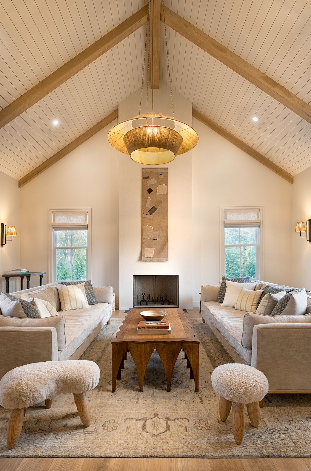 Living room with vaulted ceiling, two sofas with cushions, a wooden table, a fireplace, and window views of greenery.