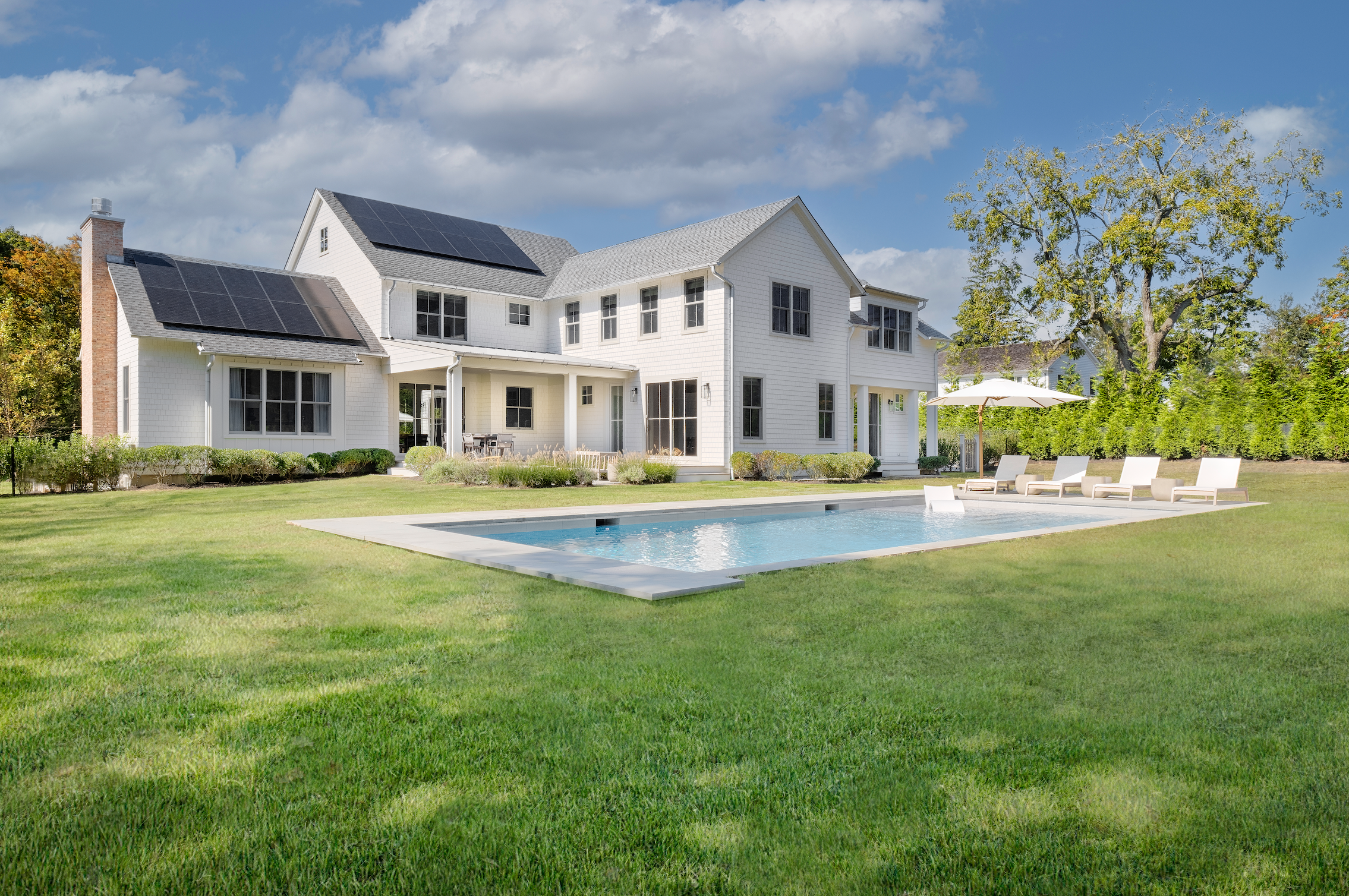 A large suburban house with white exterior walls and multiple gables, some with solar panels, sitting on a well-manicured grassy lawn. In front of the house is a rectangular swimming pool with lounge chairs and an umbrella on the patio. A large tree and bushes encircle the yard, with a partly cloudy sky overhead.