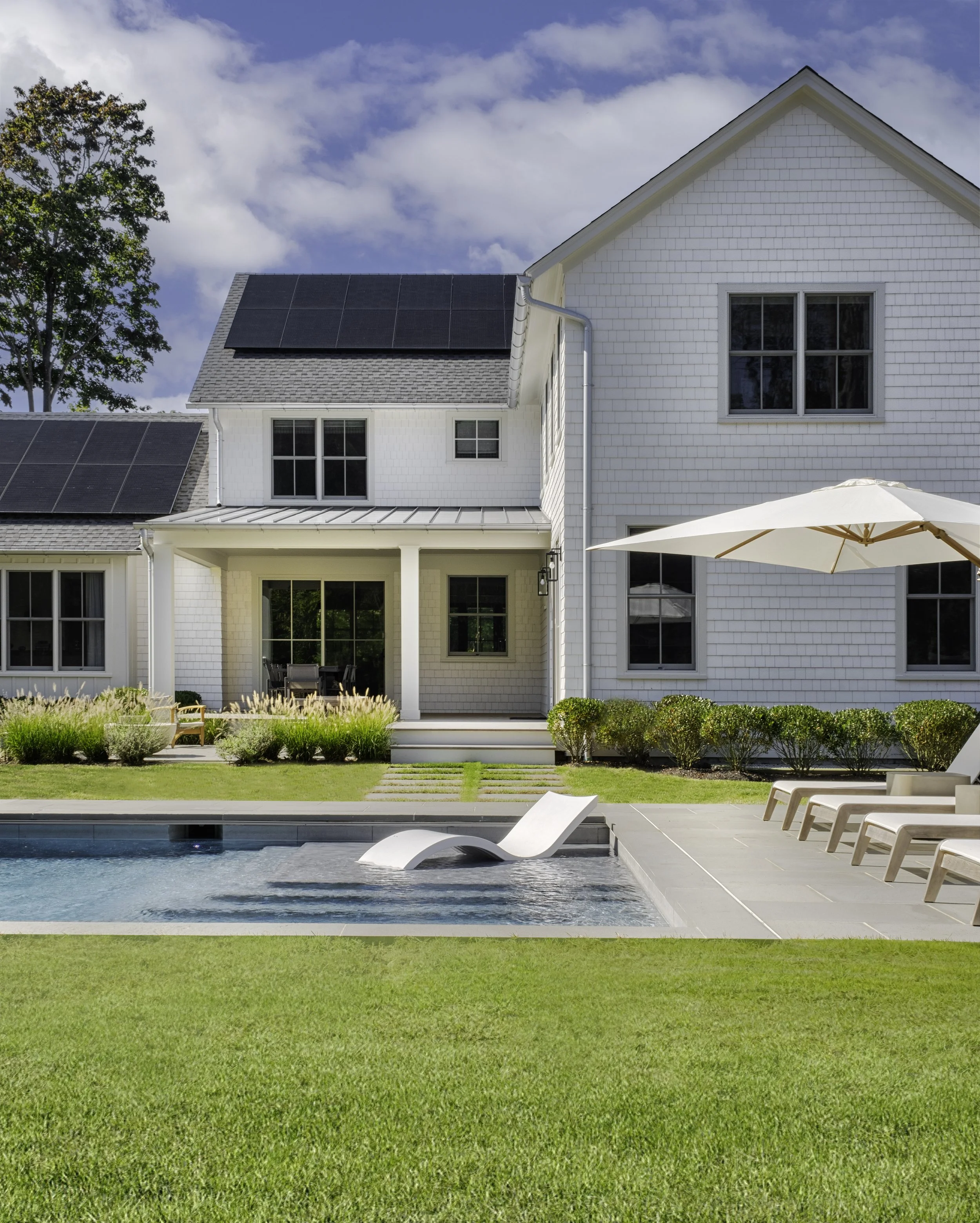 White house with black solar panels on roof, backyard with swimming pool, lounge chairs, umbrella, and manicured lawn.