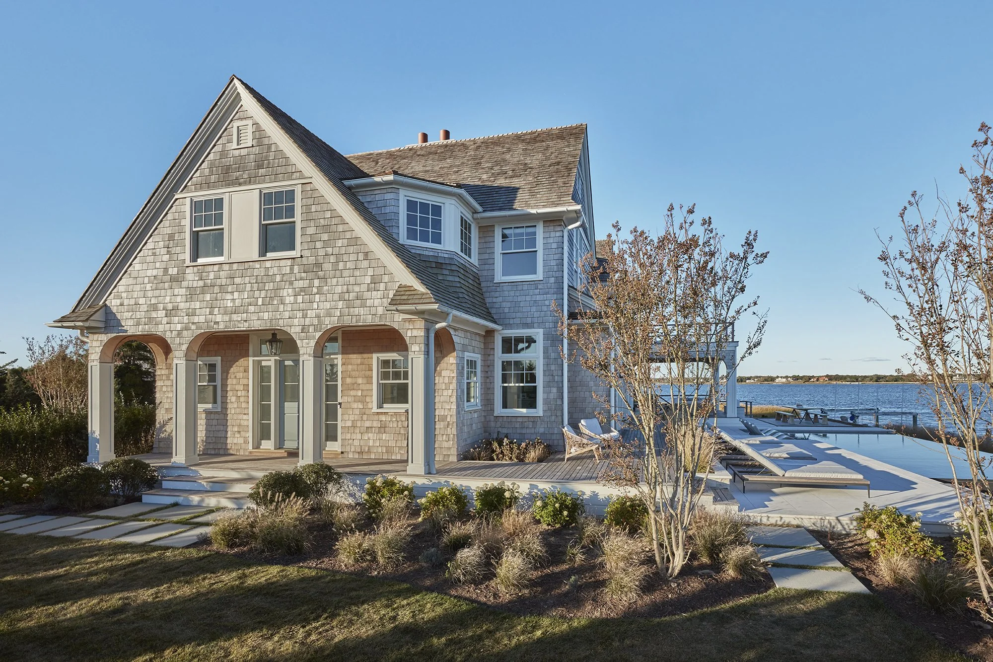 A large coastal house with gray shingle siding, multiple windows, a covered porch, and a deck with lounge chairs overlooking water, under a clear blue sky.
