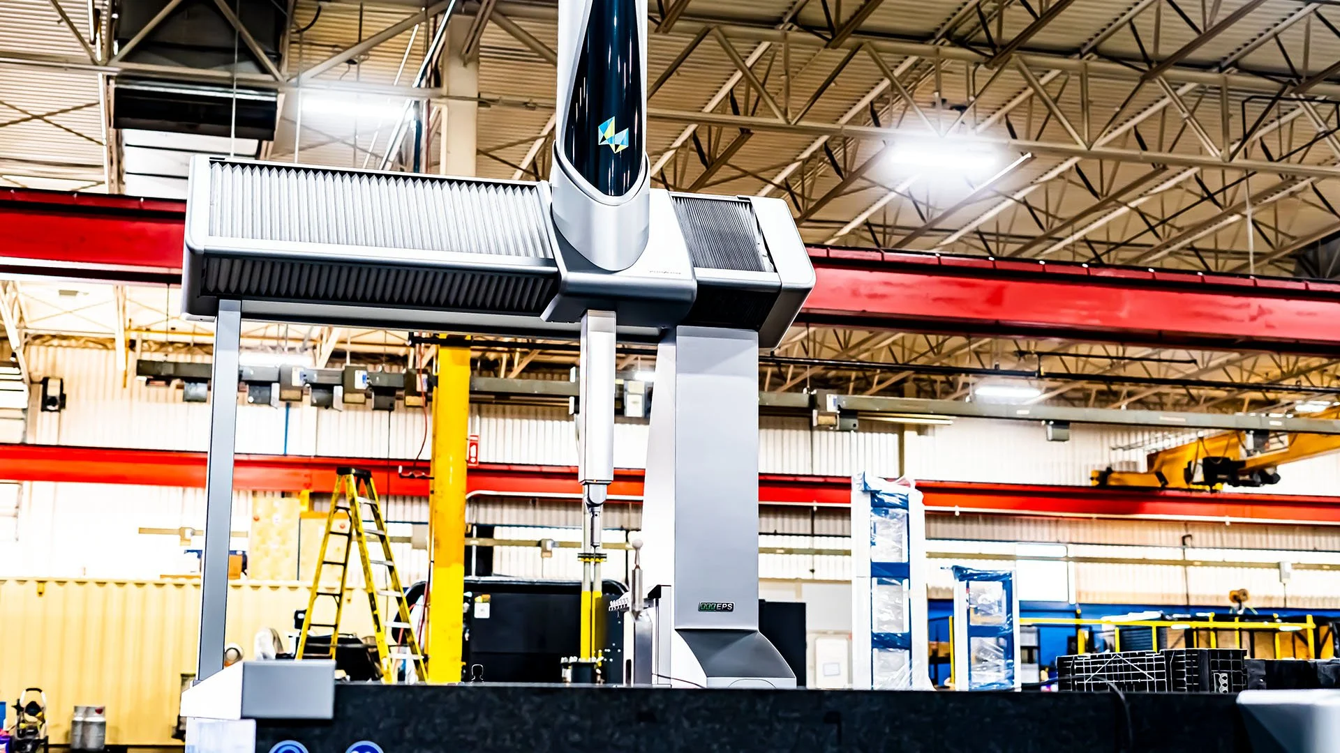 Industrial manufacturing robot in a factory with yellow ladder, overhead cranes, and metal structures in the background.