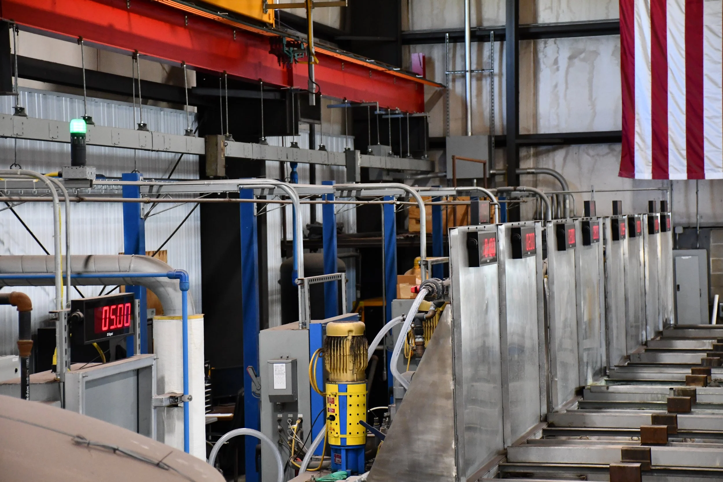 Industrial factory setup with metal machinery, pipes, and digital displays, including a partially visible American flag on the right.