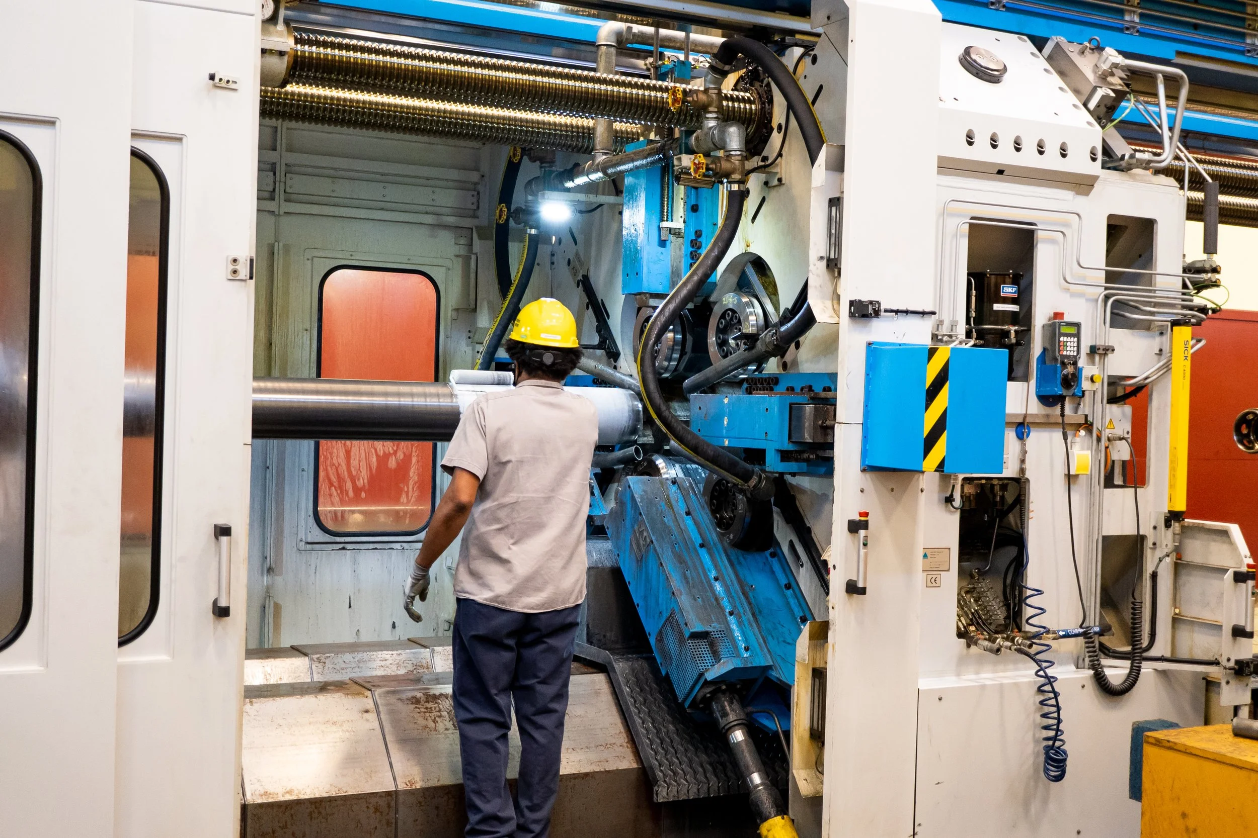 A worker in a yellow safety helmet and work clothes operating large industrial machinery in a factory or manufacturing plant.