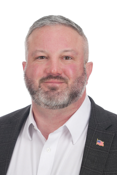 A middle-aged man with short gray hair and a beard, wearing a white shirt and a dark blazer with a small American flag pin on the lapel, smiling against a plain white background.