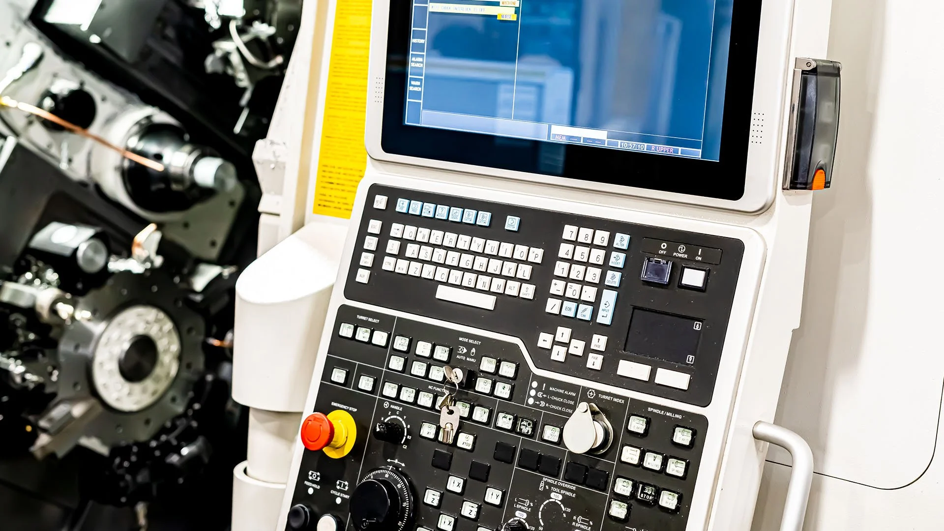 Close-up of a CNC machine control panel with a screen, numerous buttons, and switches, with mechanical parts of the machine visible in the background.