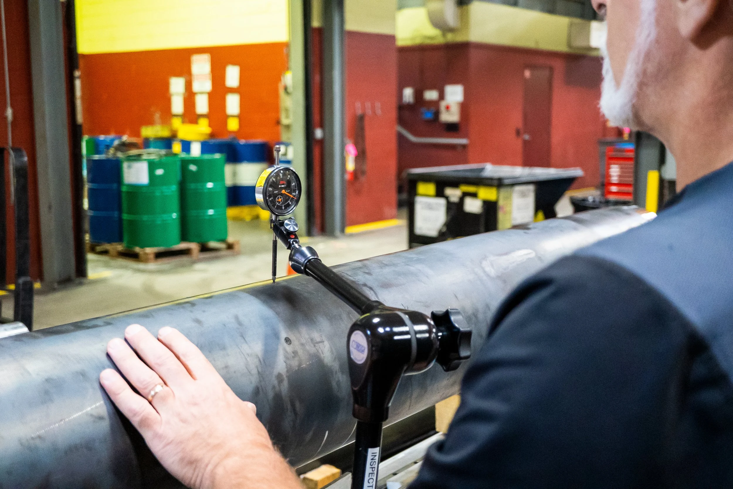 A man inspects a large cylindrical pipe in an industrial setting, using a dial gauge attached to a height measurement tool. In the background, there are pallets of colored drums and various industrial equipment.