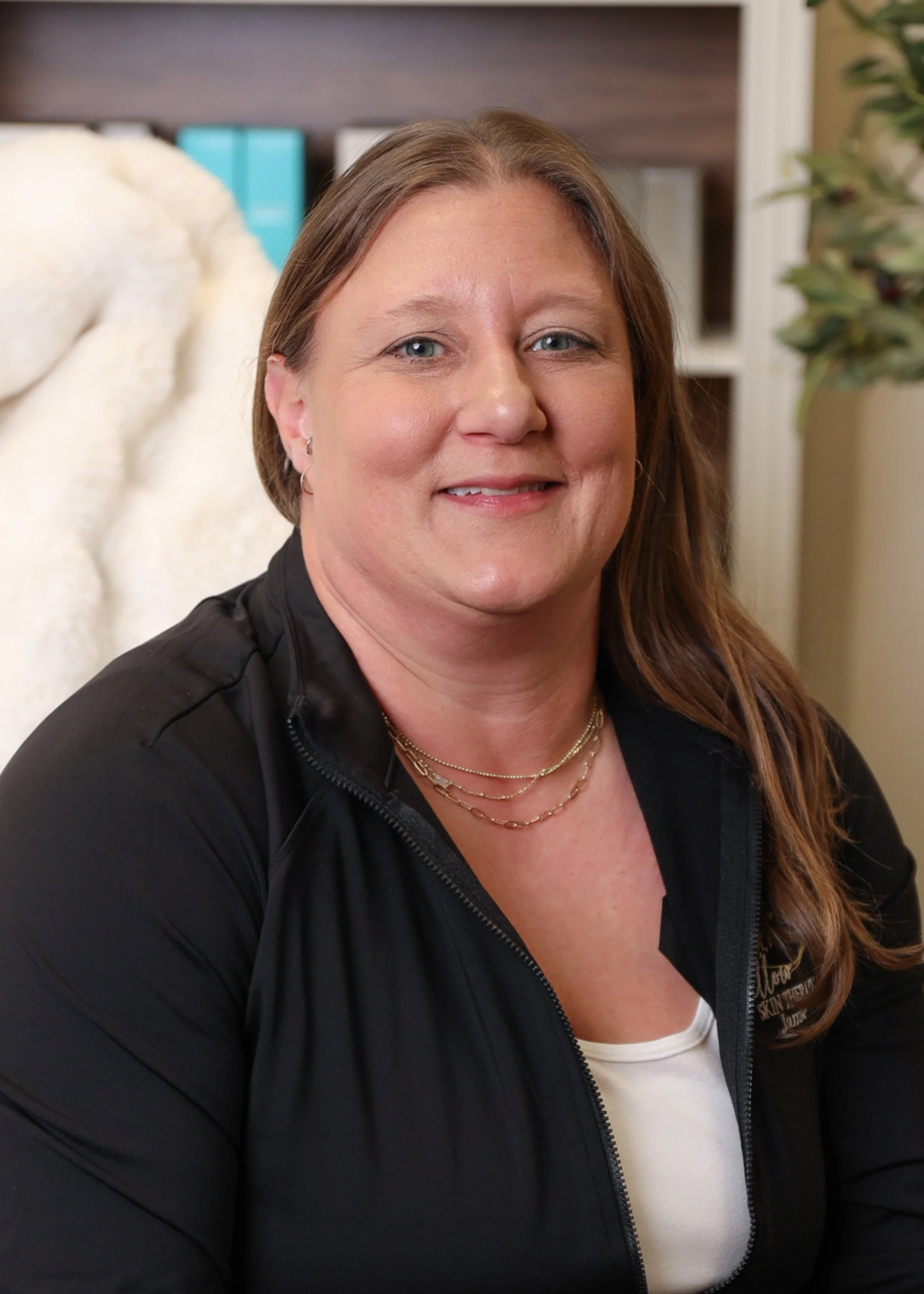 A woman with light brown hair, wearing a black zip-up jacket over a white top, sitting in front of a bookshelf and smiling at the camera.