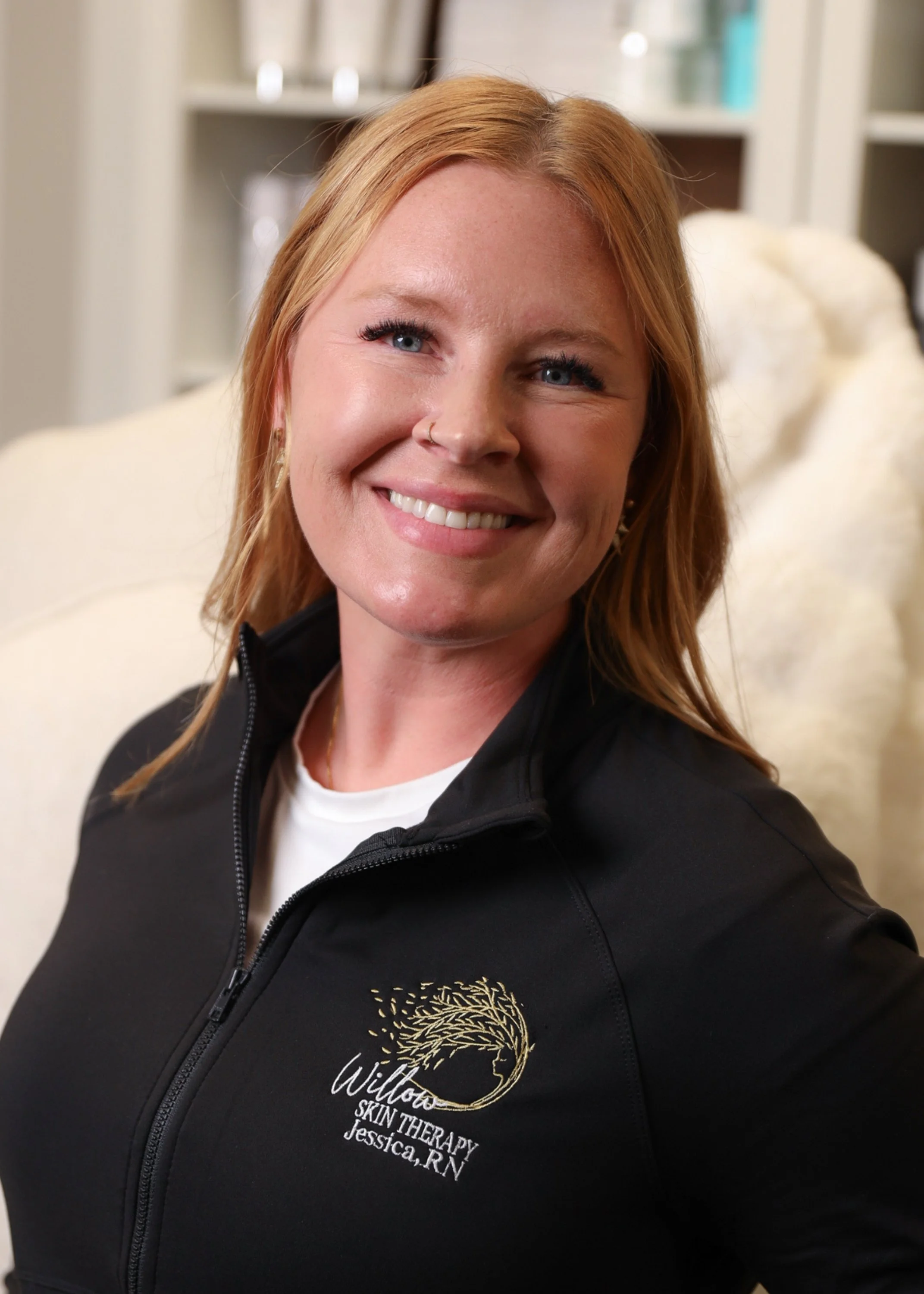 A woman with red hair smiling at the camera, wearing a black jacket with a logo that says 'Wilto Skin Therapy Jessica, RN', sitting on a beige couch in a room with shelves in the background.