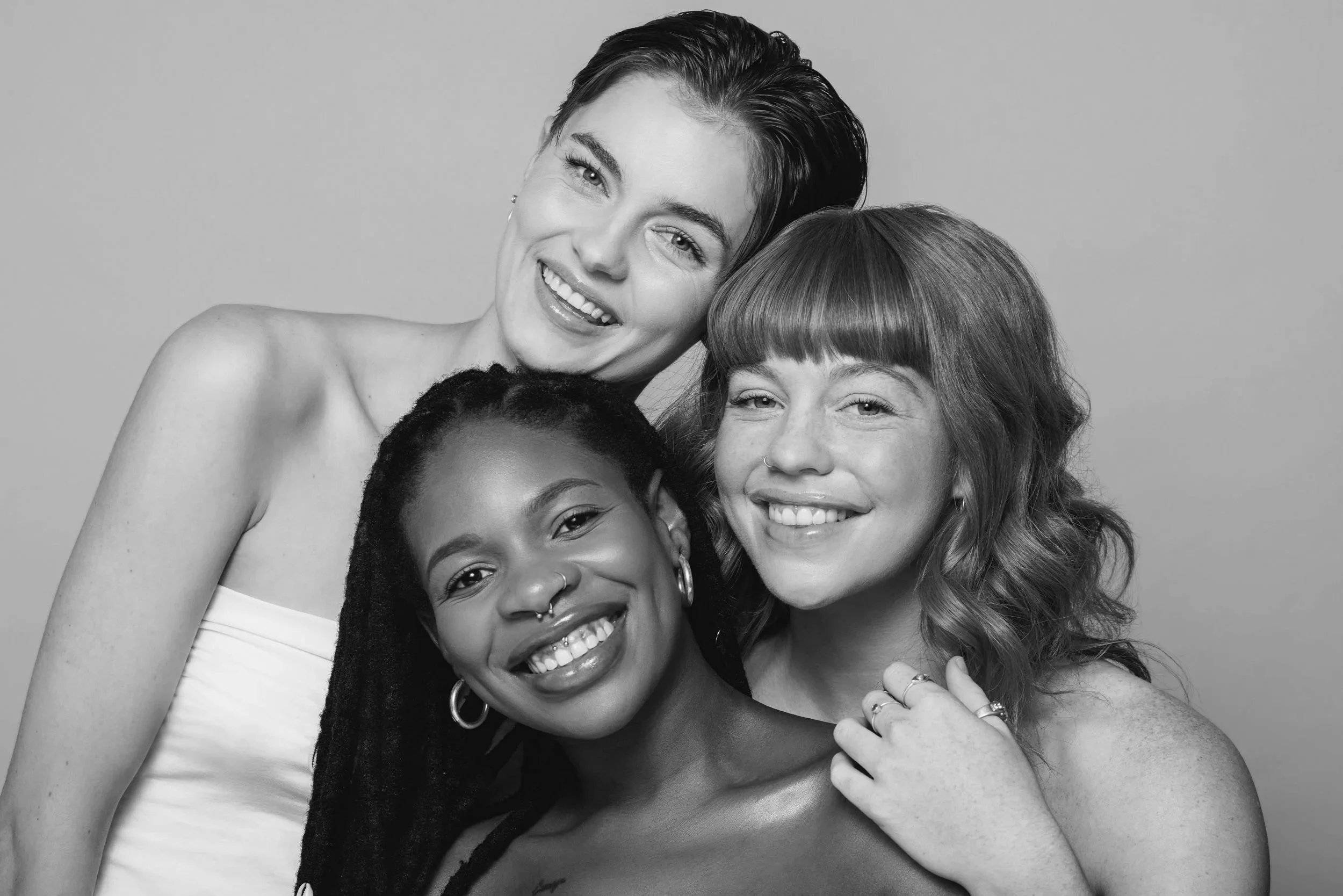 Three smiling women of diverse ethnicities posing together in a black-and-white portrait.