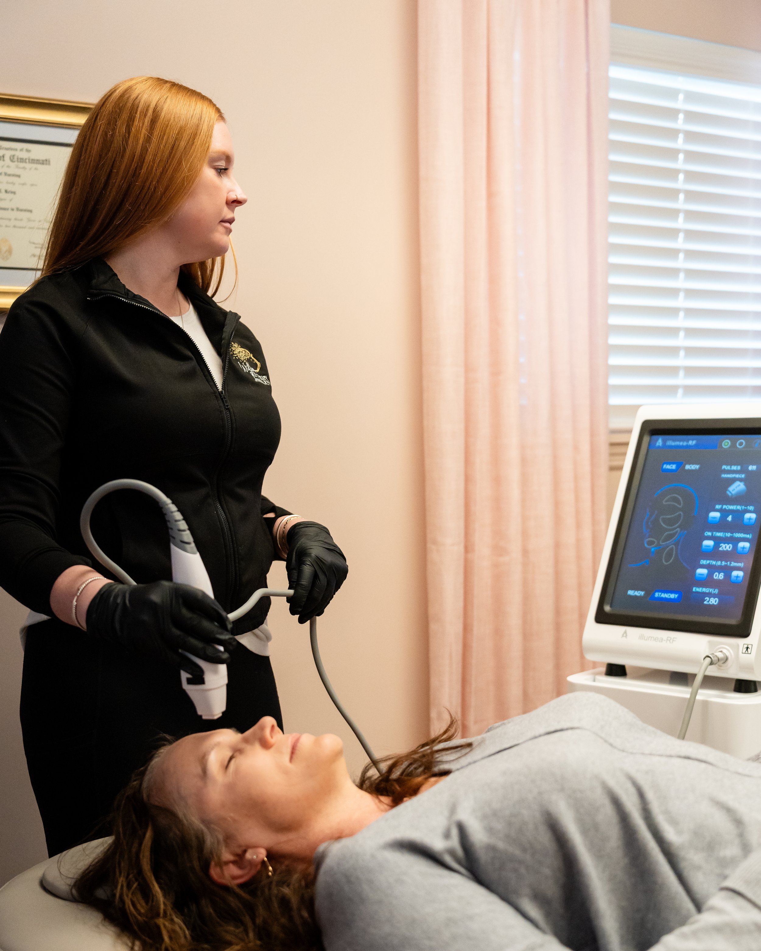 A Cincinnati Med Spa medical professional wearing black gloves and a black zip-up jacket performing an ultrasound scan on a woman lying on a medical bed, with a ultrasound machine nearby and a framed certificate on the wall.