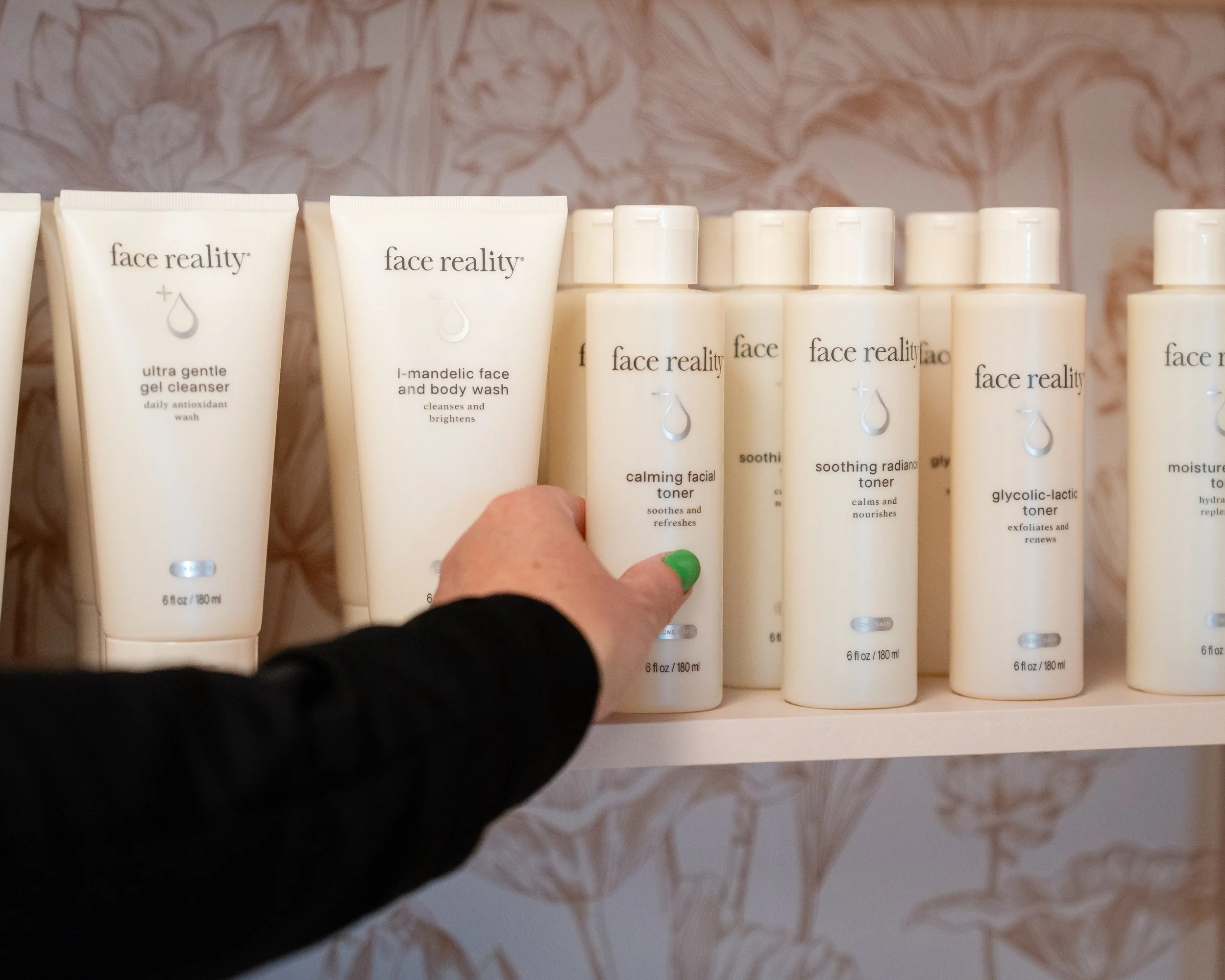 A person is selecting a calming facial toner from a shelf of skincare products. The shelf displays multiple bottles and tubes of Face Reality skincare products, including gentle gel cleanser, mandelic face and body wash, calming facial toner, soothing radiance toner, and glycol-lactic toner, against a floral patterned background.