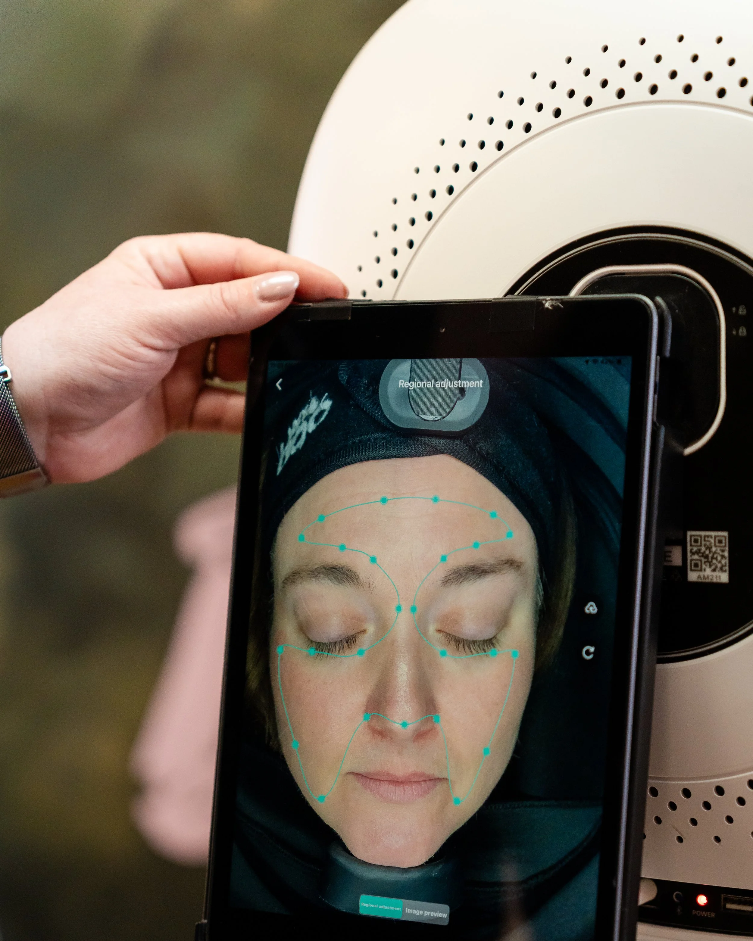 A person using a tablet to scan a person's face for facial recognition or analysis, with facial mapping points visible on the screen, while the person lies down with eyes closed.