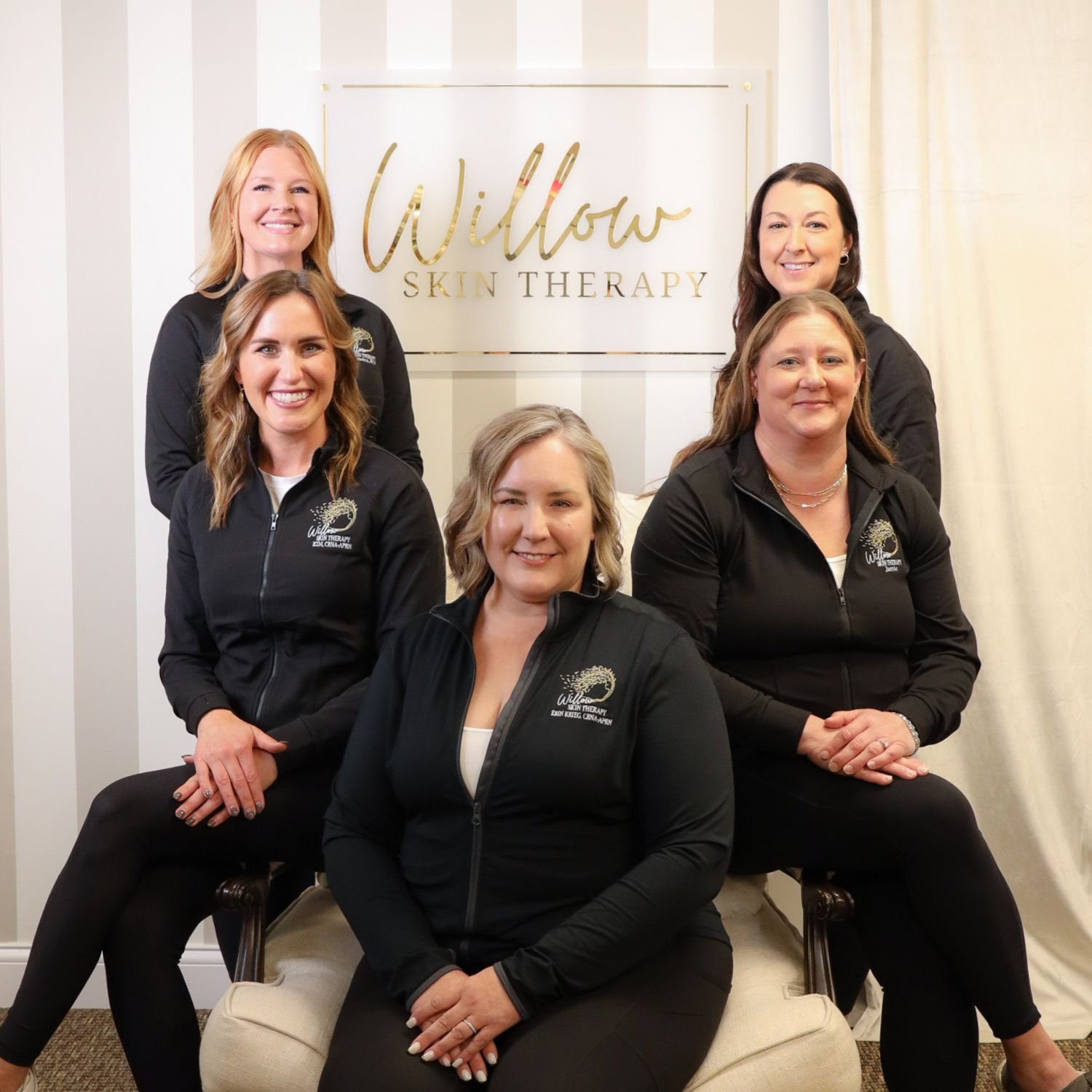 Group of five women wearing black jackets with 'Willow Skin Therapy' logo, sitting and standing in front of a sign that reads 'Willow Skin Therapy.'