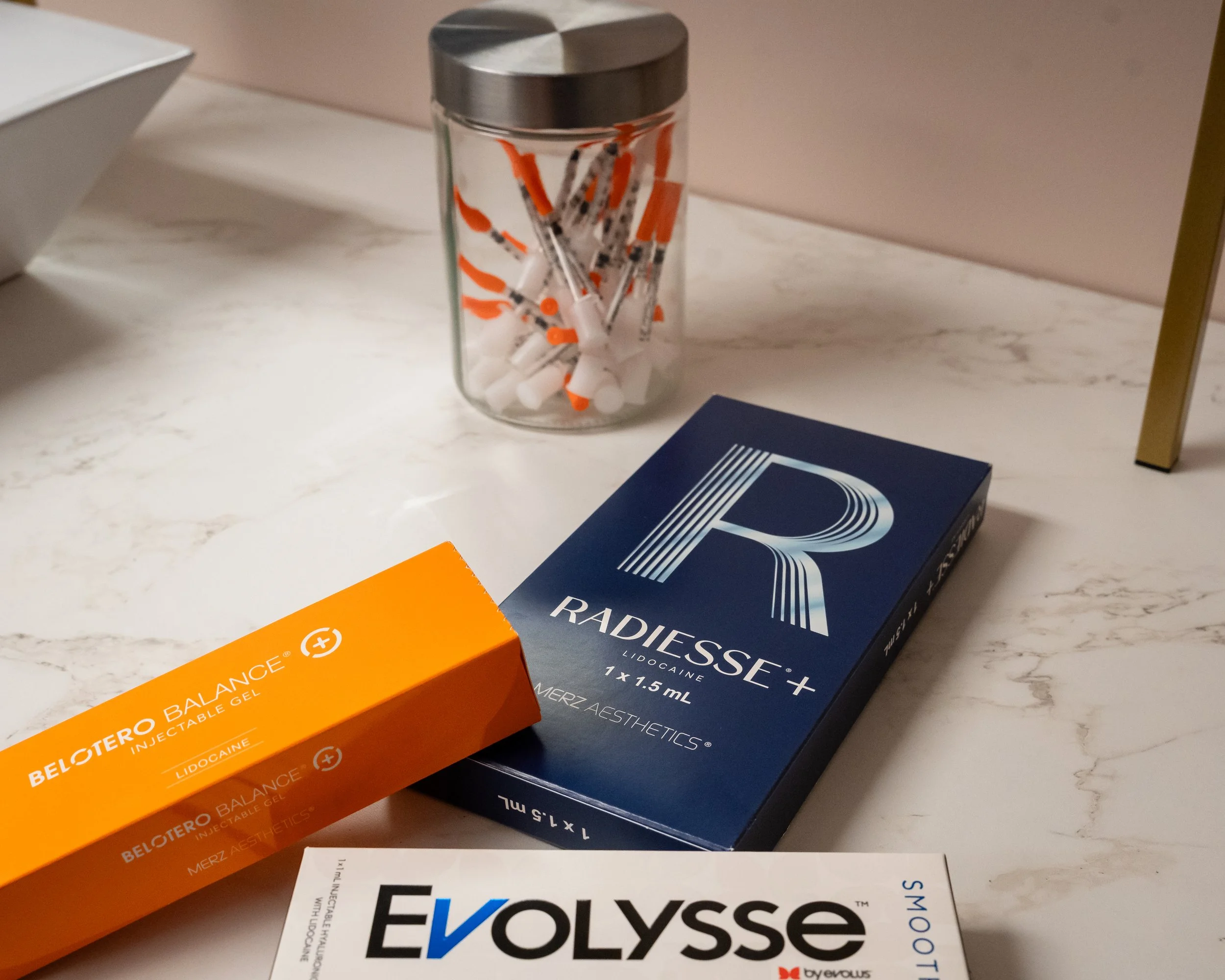 A marble countertop with a jar of syringes, a box of Radiesse dermal filler, a box of Belotero Balance dermal filler, and a box of Evolyse skincare product.