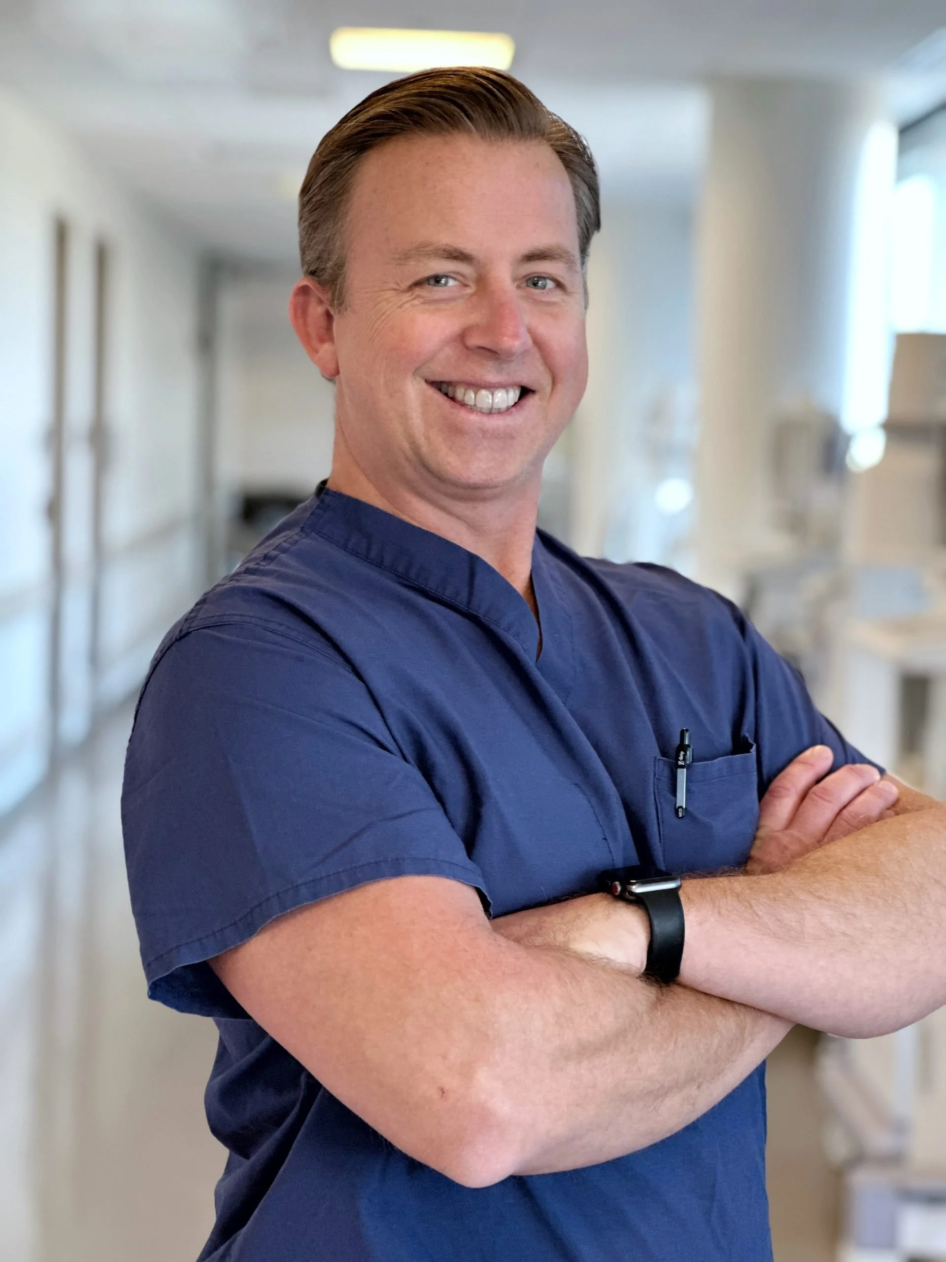 A male healthcare professional in navy scrubs smiling with arms crossed in a hospital corridor.