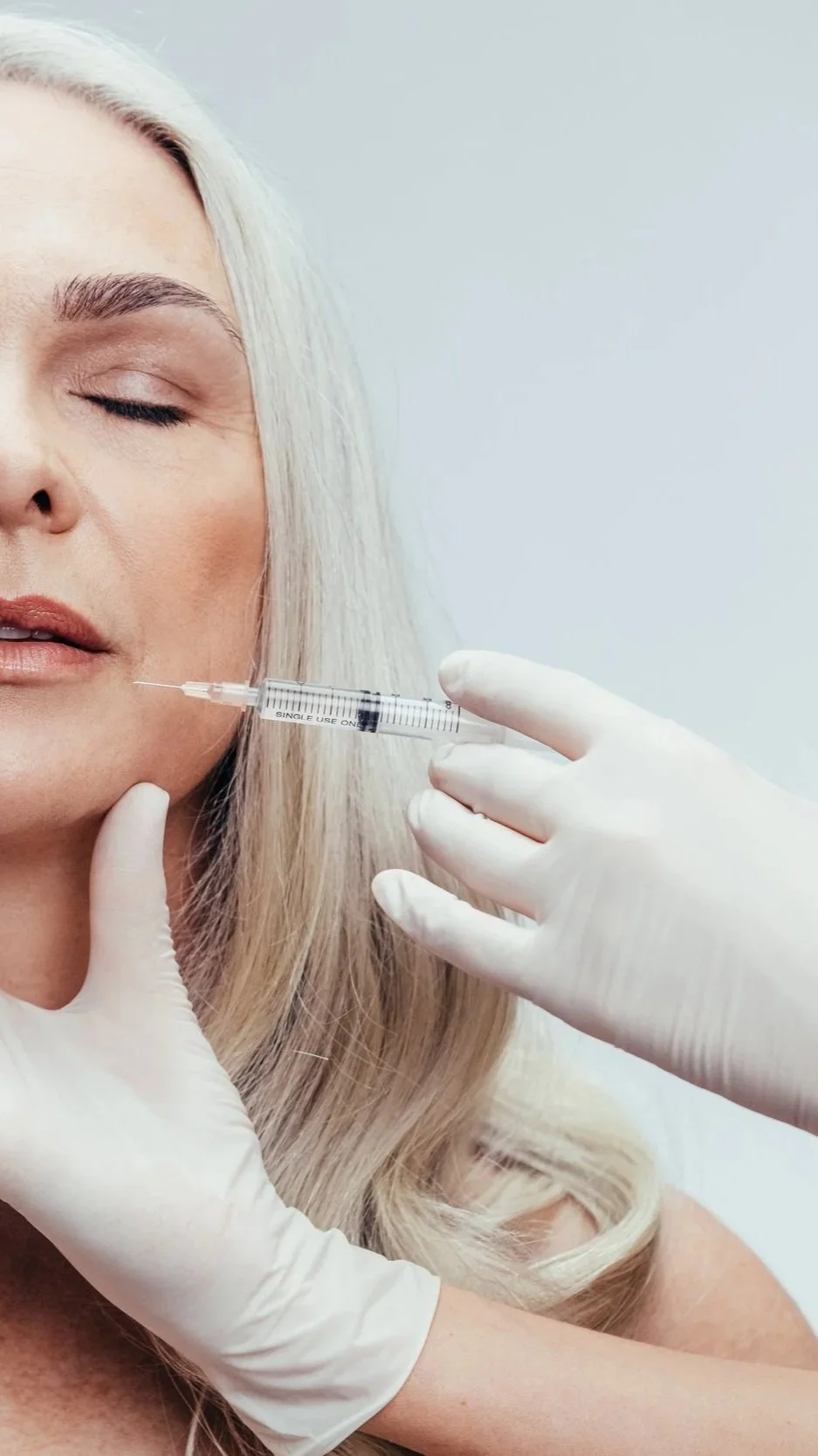 A woman with blonde hair receiving a cosmetic injection in her cheek from a syringe held by a gloved hand.