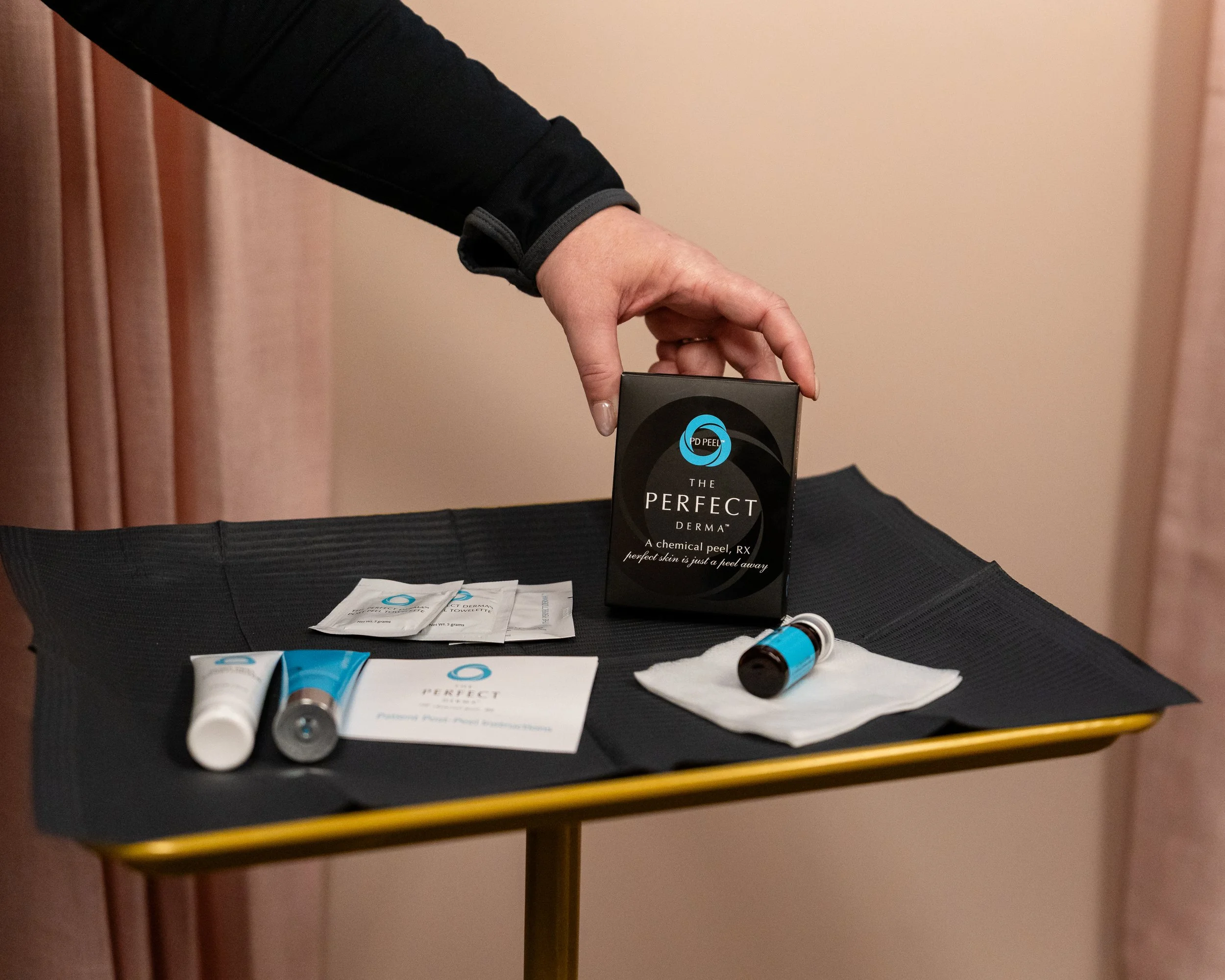 A person arranging skincare products on a black table, including a box labeled 'The Perfect Derma' chemical peel, small vials, tubes, and sample packets. The background features pink curtains.