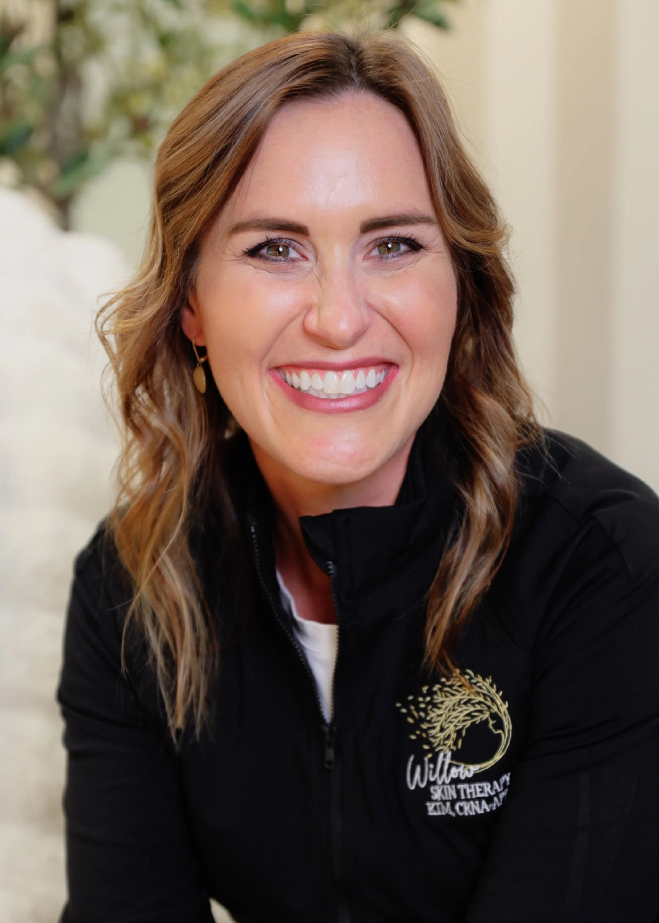 A woman with wavy, shoulder-length brown hair and light skin, smiling and wearing a black jacket with a logo that reads 'Willow Skin Therapy' along with a tree graphic, in an indoor setting with plants in the background.
