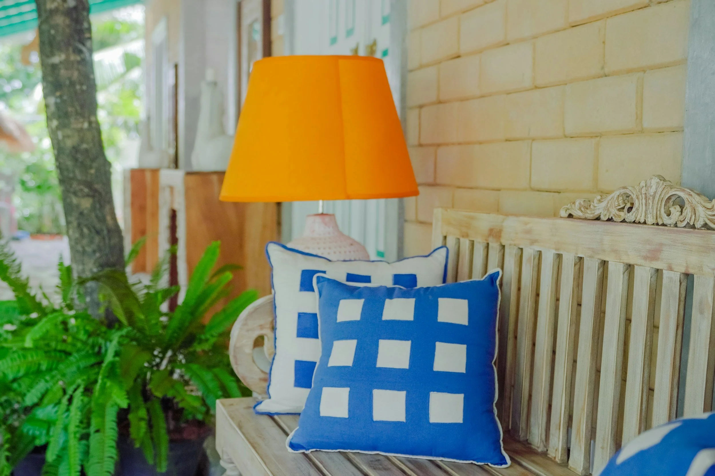 A cozy corner with a wooden bench adorned with two blue and white checkered pillows, a yellow lampshade on a table, a lush green fern plant, and natural wood and brick wall accents.